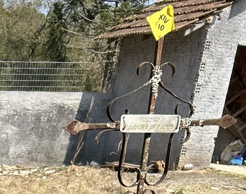 Rusty metal cross with a plaque inscribed 'Jaskowski' in front of a weathered stone wall and a corrugated metal roof structure.