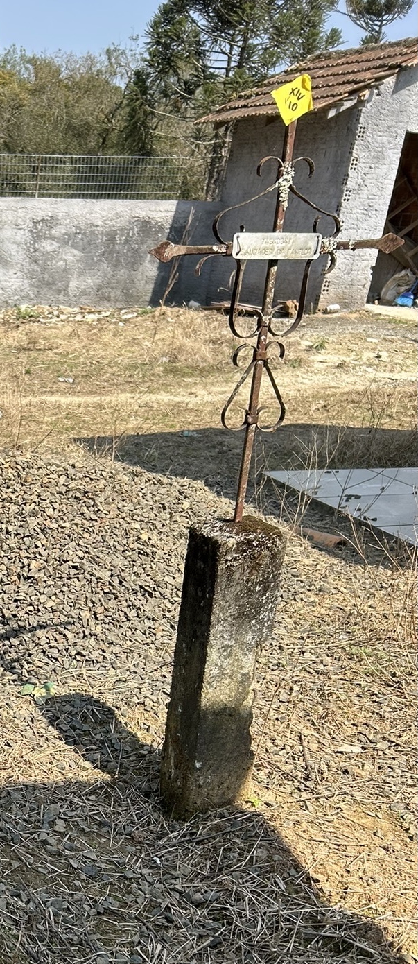 Rusted metal cross on a stone base in a grassy area with a small building in the background.