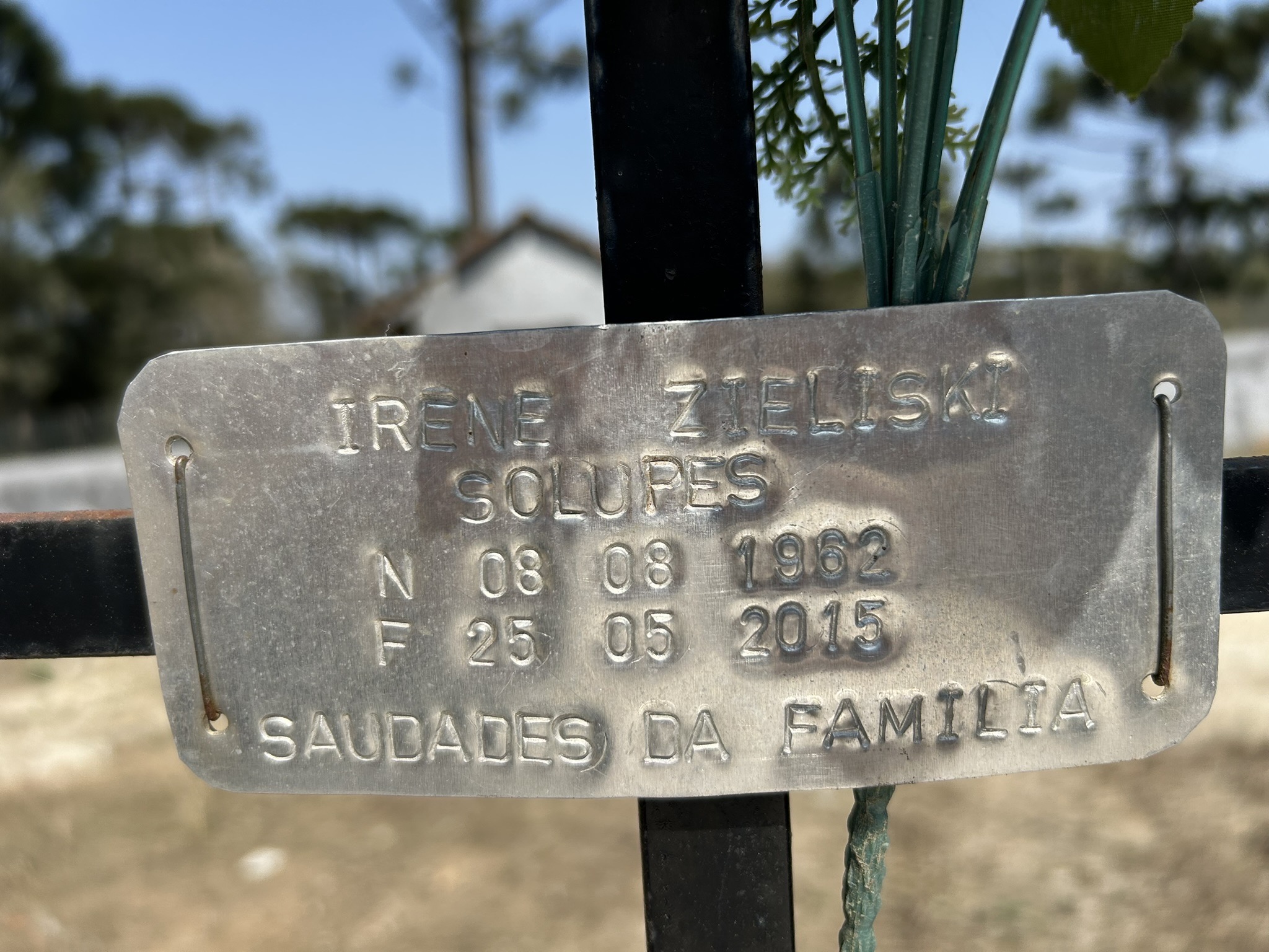 Metal plaque on grave with engraved text: Irene Zieliski, Solupes, N 08 08 1962, F 25 05 2015, Saudades da Familia. In the background, blurred trees and a building.