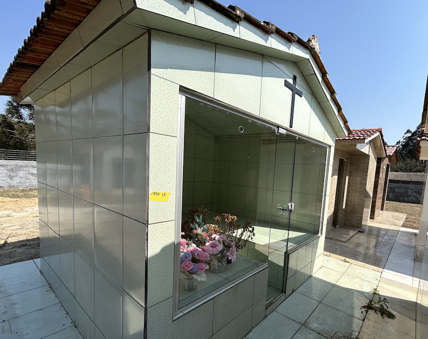 A small chapel with a tiled façade and glass front, incorporating floral compositions. A cross is mounted on the roof. The chapel is part of a series of similar structures.