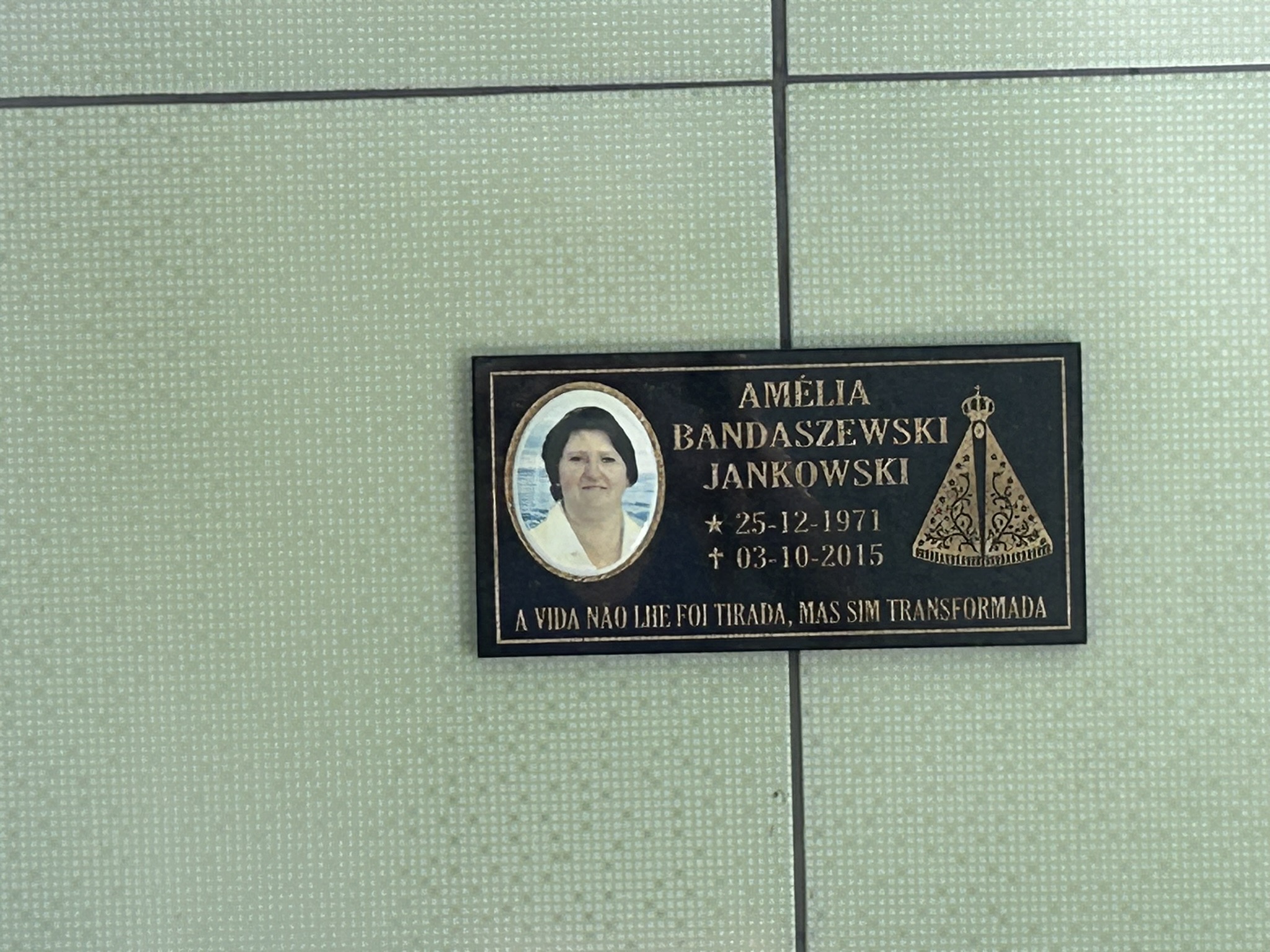 Plaque with a photograph of a woman and text: 'Amélia Bandaszewski Jankowski, 25-12-1971, 03-10-2015'. Inscription in Portuguese: 'A vida não lhe foi tirada, mas sim transformada'.