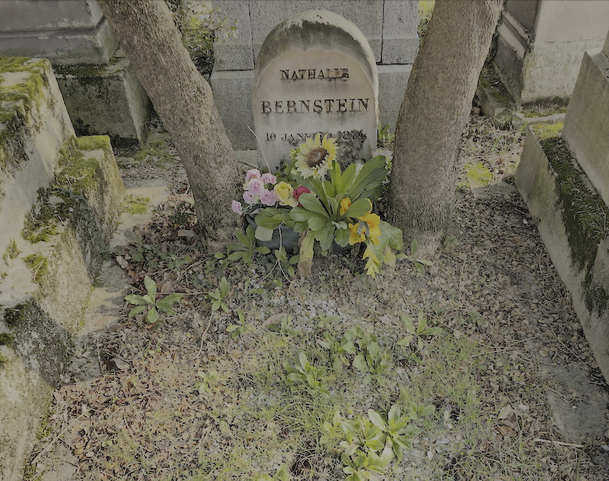 Pierre tombale de Nathalie Bernstein avec un sommet semi-circulaire, inscrite avec son nom et sa date. Entourée d'arbres et de fleurs, la pierre est enfoncée dans le sol.