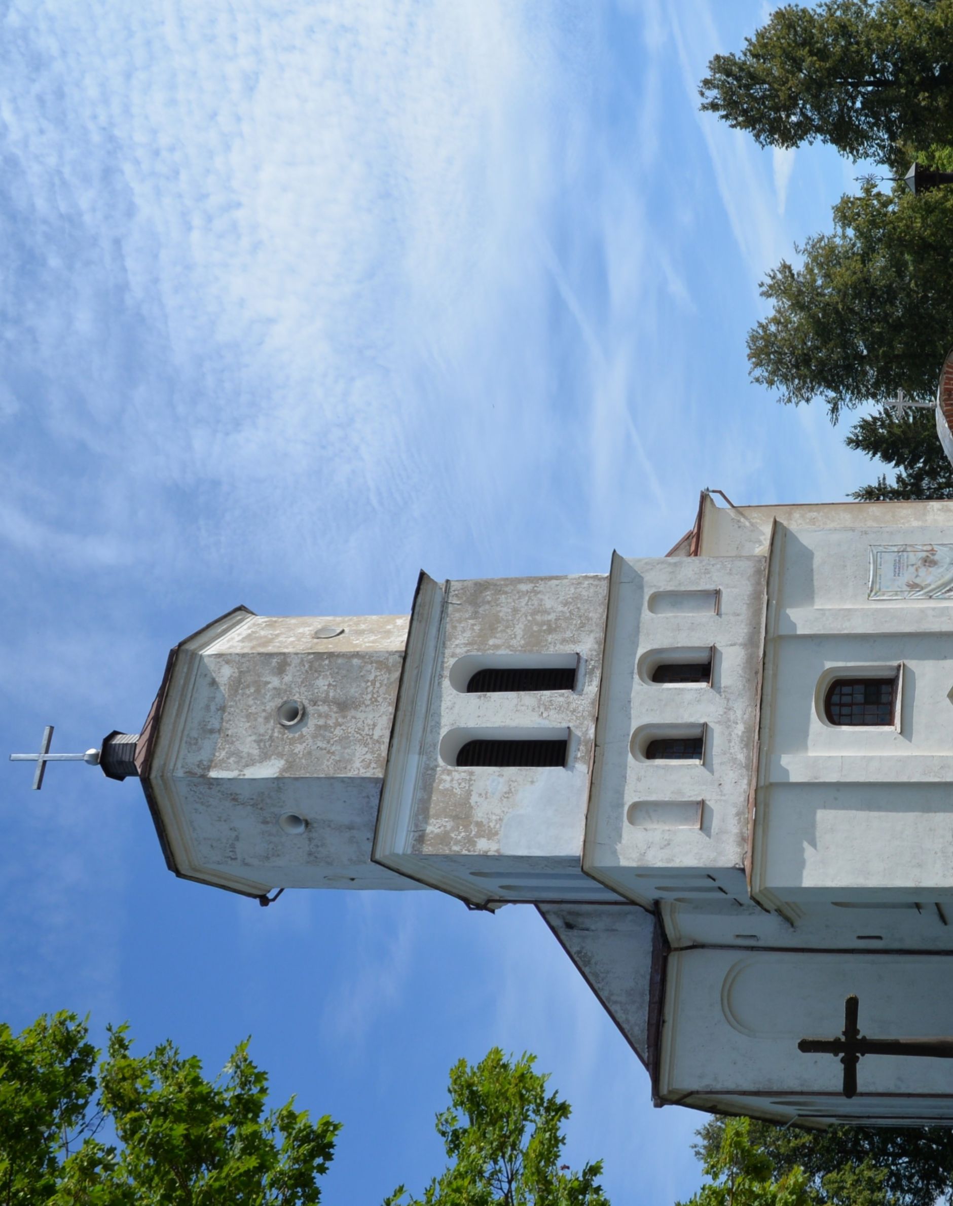 The Church in Siesikai with a massive, austere structure featuring a four-story tower with a cross on top. The facade has arched windows and a simple decoration. Trees and a clear sky in the background.