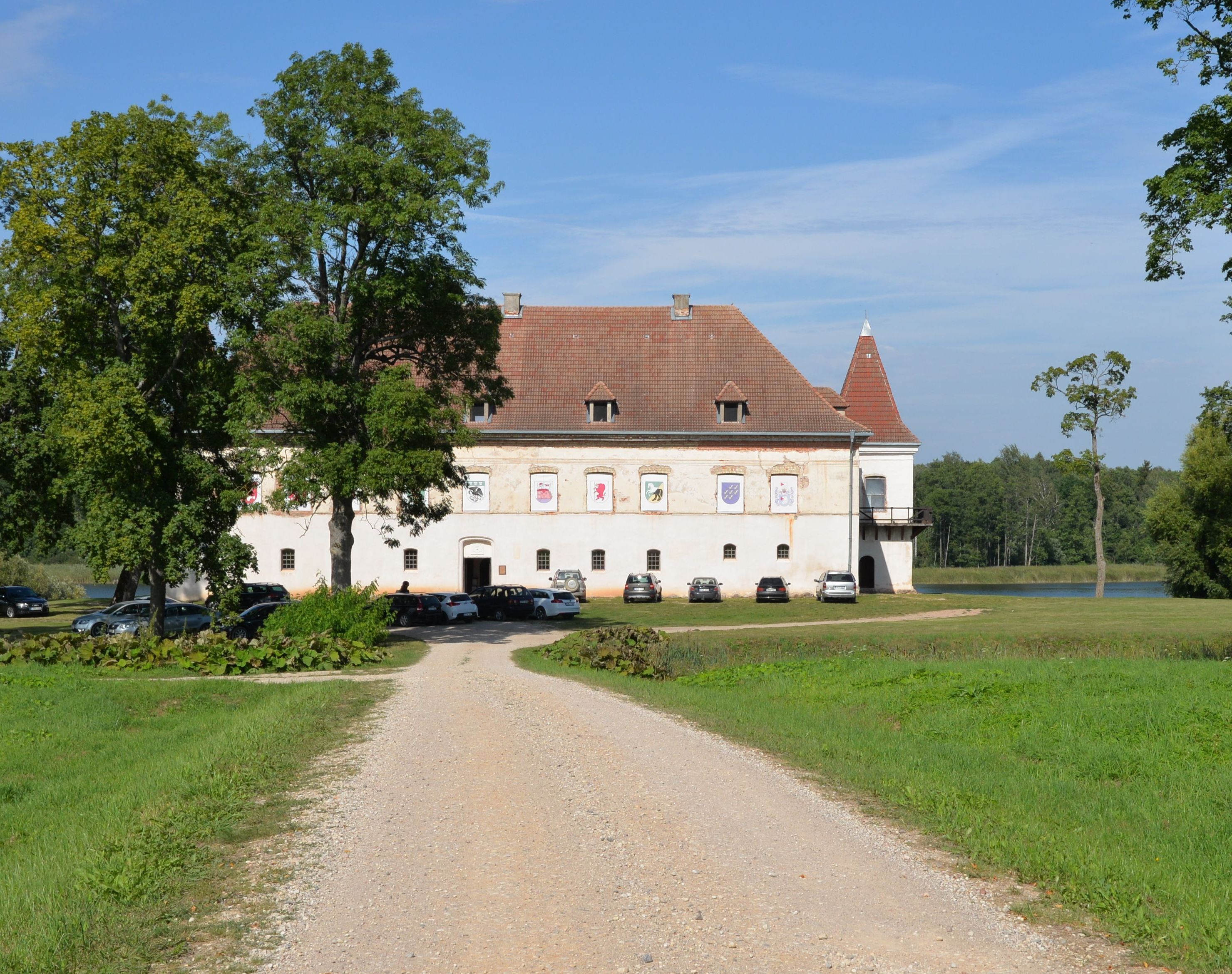Siesikai Castle with a red-tiled roof and white walls, surrounded by trees and a gravel path leading to the entrance. Cars are parked in front of the building. A lake is visible in the background.