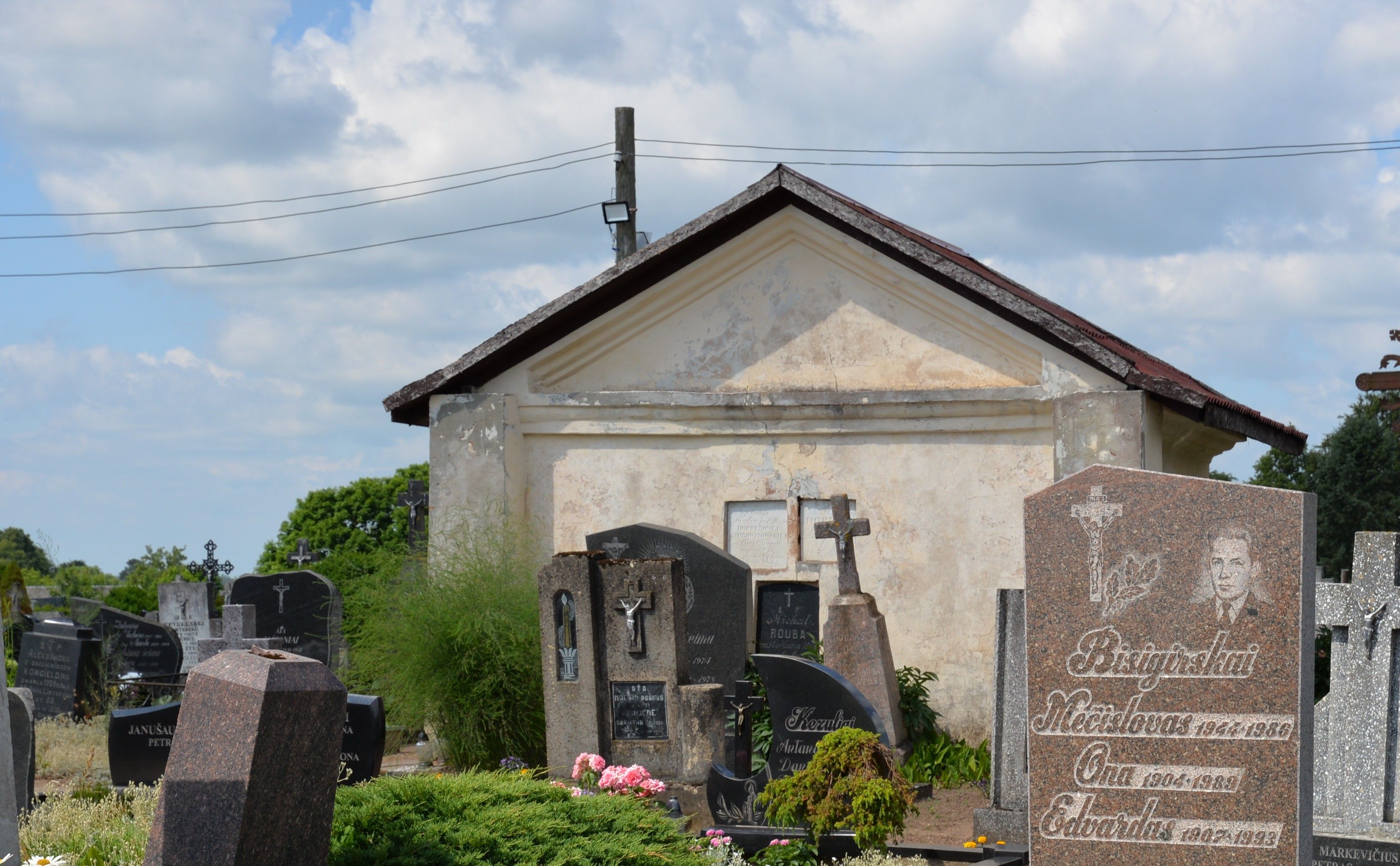 Gravestones and a chapel on the cemetery in Šėta. The chapel has a simple rectangular shape with a gabled roof. Various gravestones with inscriptions and crosses are in the foreground.