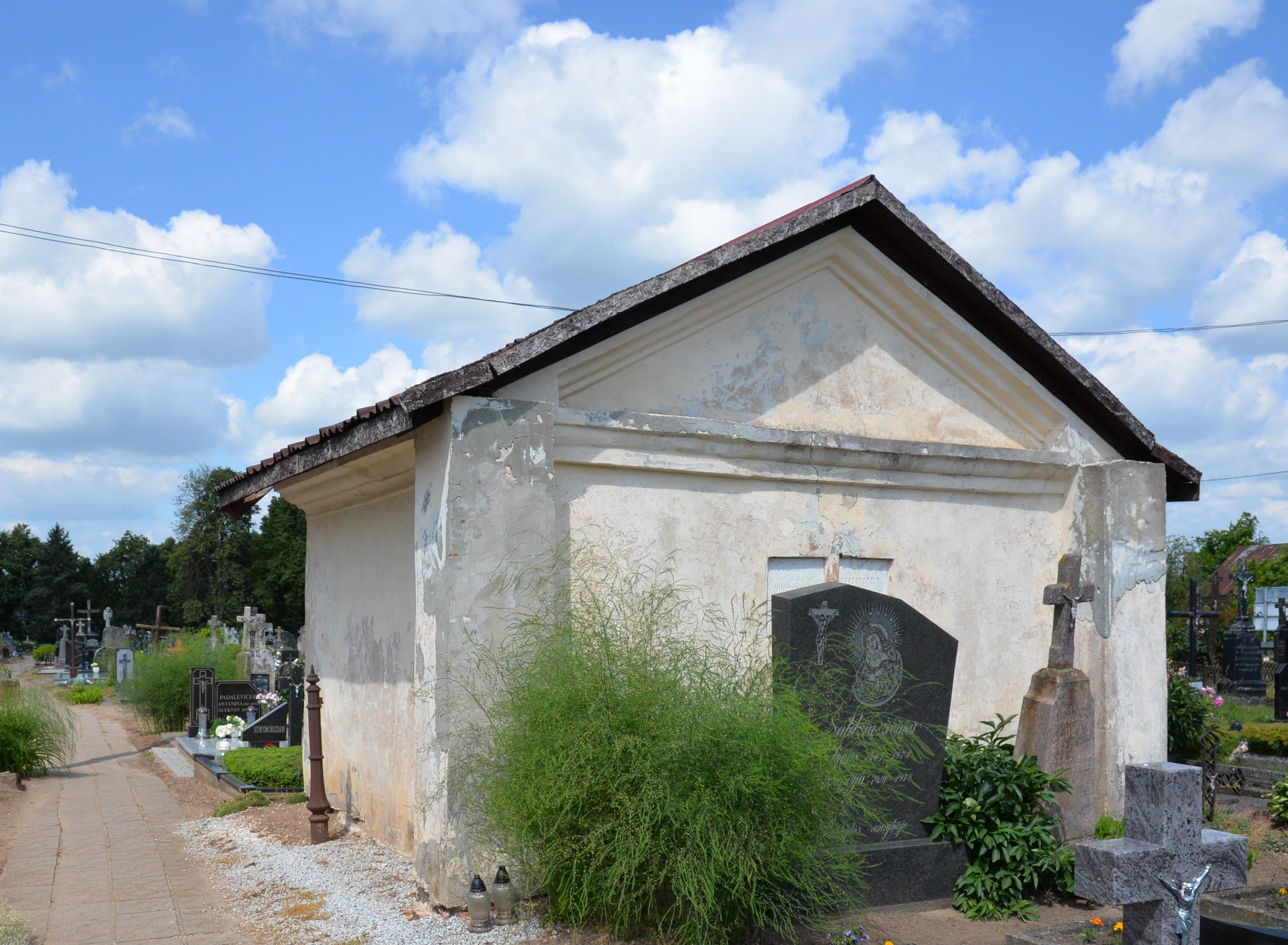 A small chapel on the cemetery in Szaty with a simple rectangular structure and a gabled roof. The facade shows signs of wear. Tombstones and crosses are visible nearby.