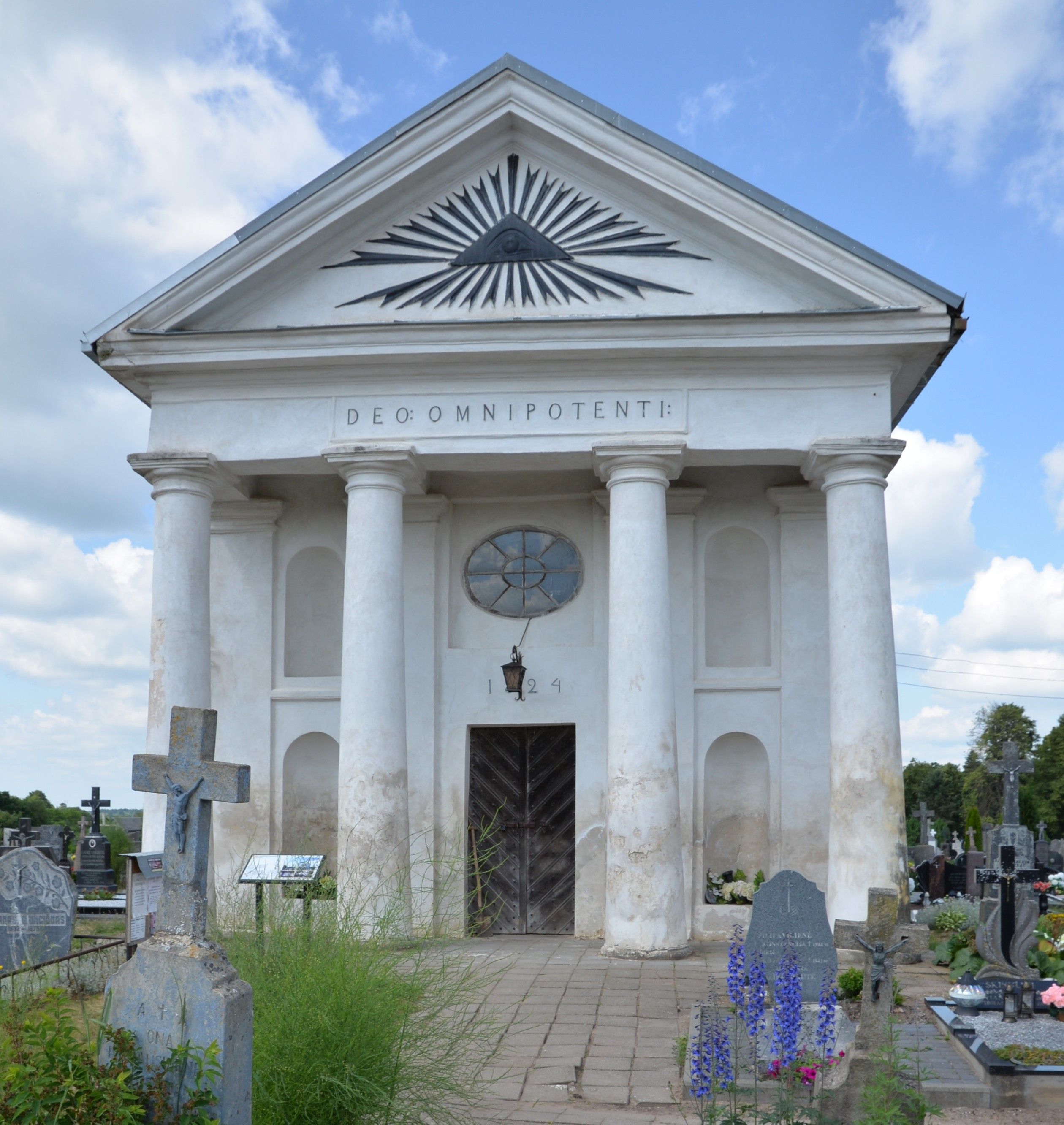 Front view of the neoclassical chapel in Szaty cemetery with four Tuscan columns, a triangular pediment featuring the Eye of Providence, and the inscription 'DEO: OMNIPOTENTI:'. The year 1824 is visible above the entrance.