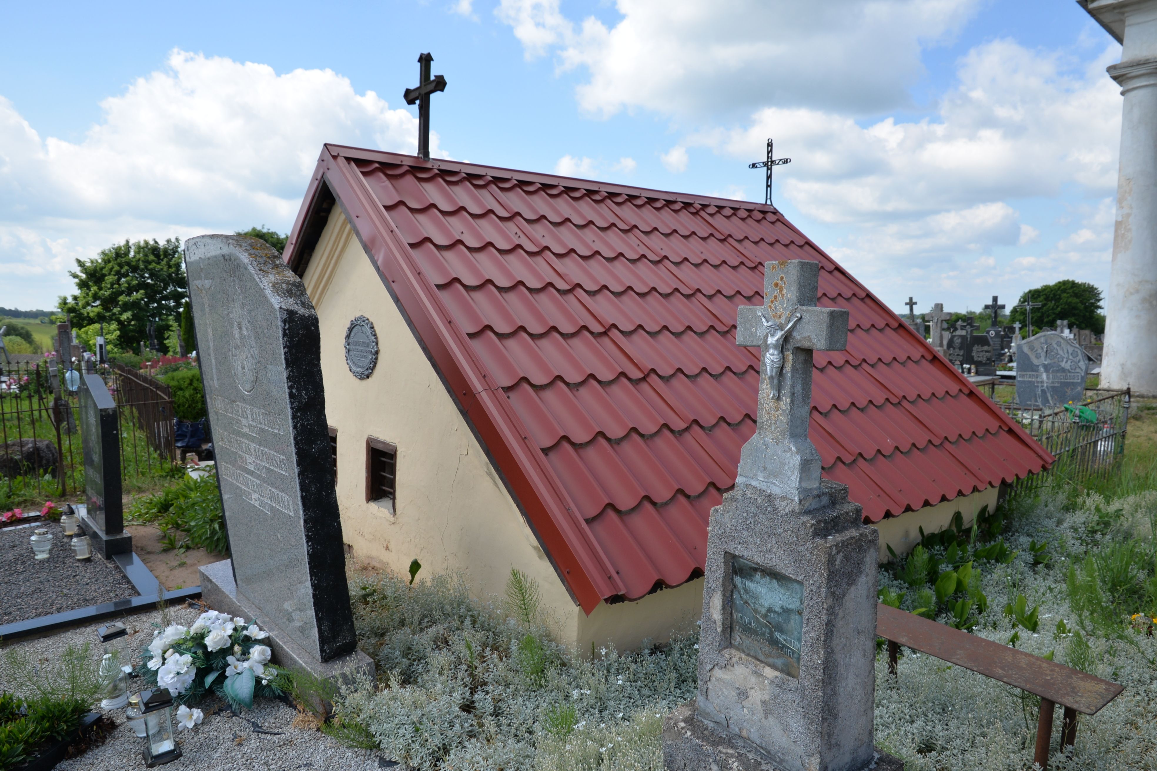 Grave with a stone cross and a small chapel with a red tiled roof at the cemetery in Szaty. The chapel has a circular plaque on its wall and is surrounded by gravestones.