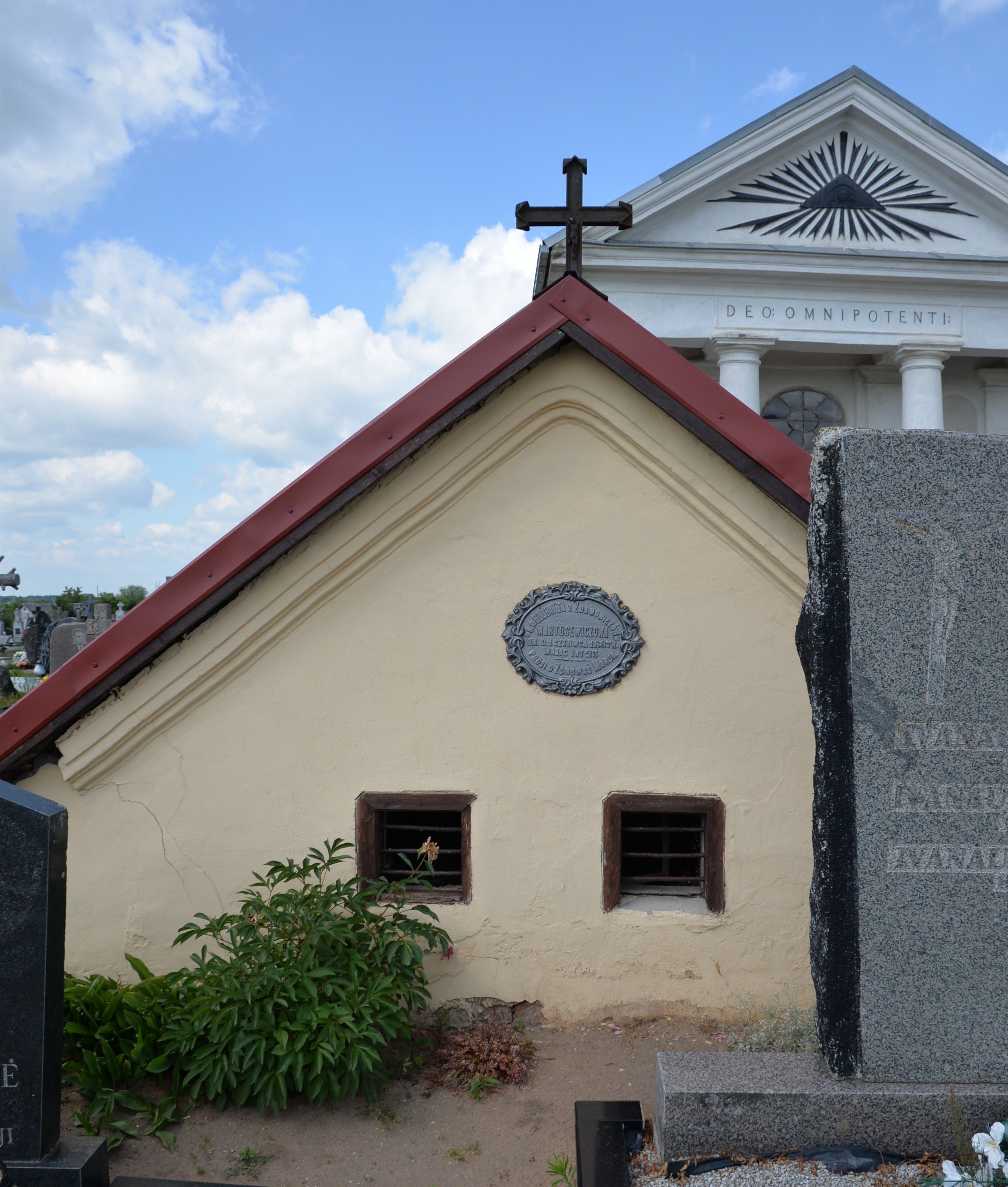 Gravestone with a cross and a plaque on a small chapel at the cemetery in Szaty. The chapel has a red gabled roof and two small barred windows. In the background, a larger building with columns and the inscription 'DEO: OMNIPOTENTI:'.