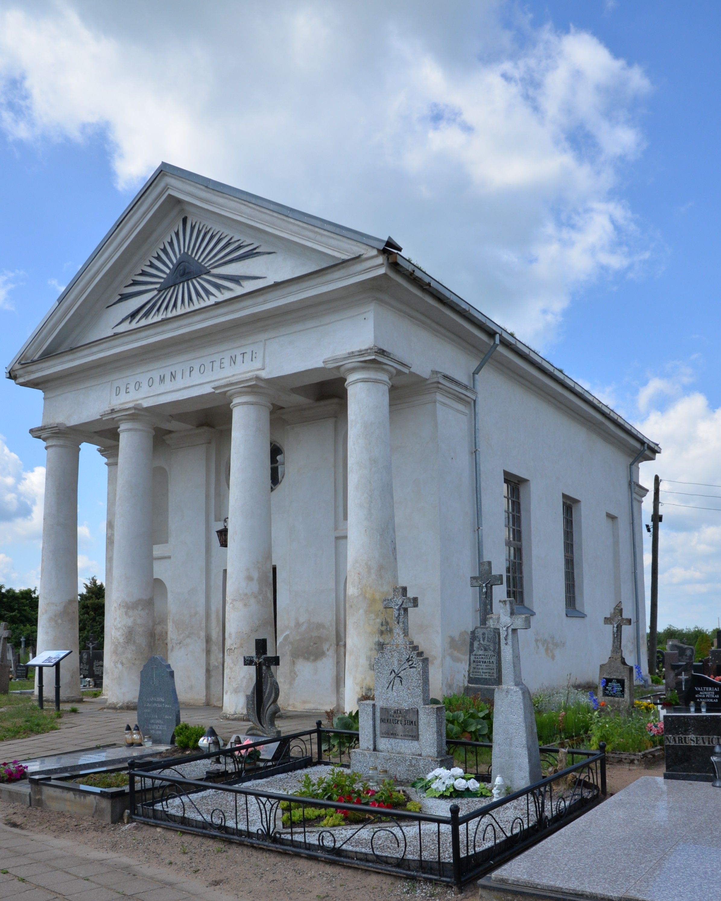 A neoclassical chapel with a four-column portico and the inscription 'DEO OMNIPOTENTI' on the pediment. Several gravestones are visible in the foreground, surrounded by flowers.