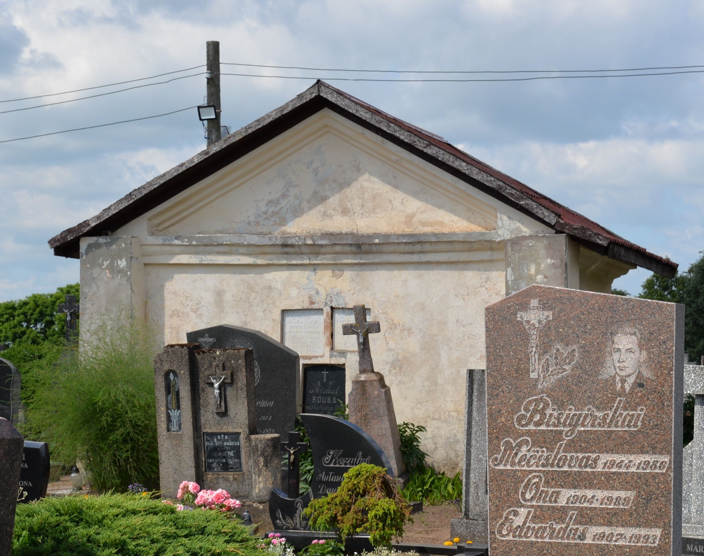 Old chapel with a gabled roof on a cemetery in Šėta, surrounded by gravestones with inscriptions and crosses. The chapel's facade shows signs of wear.