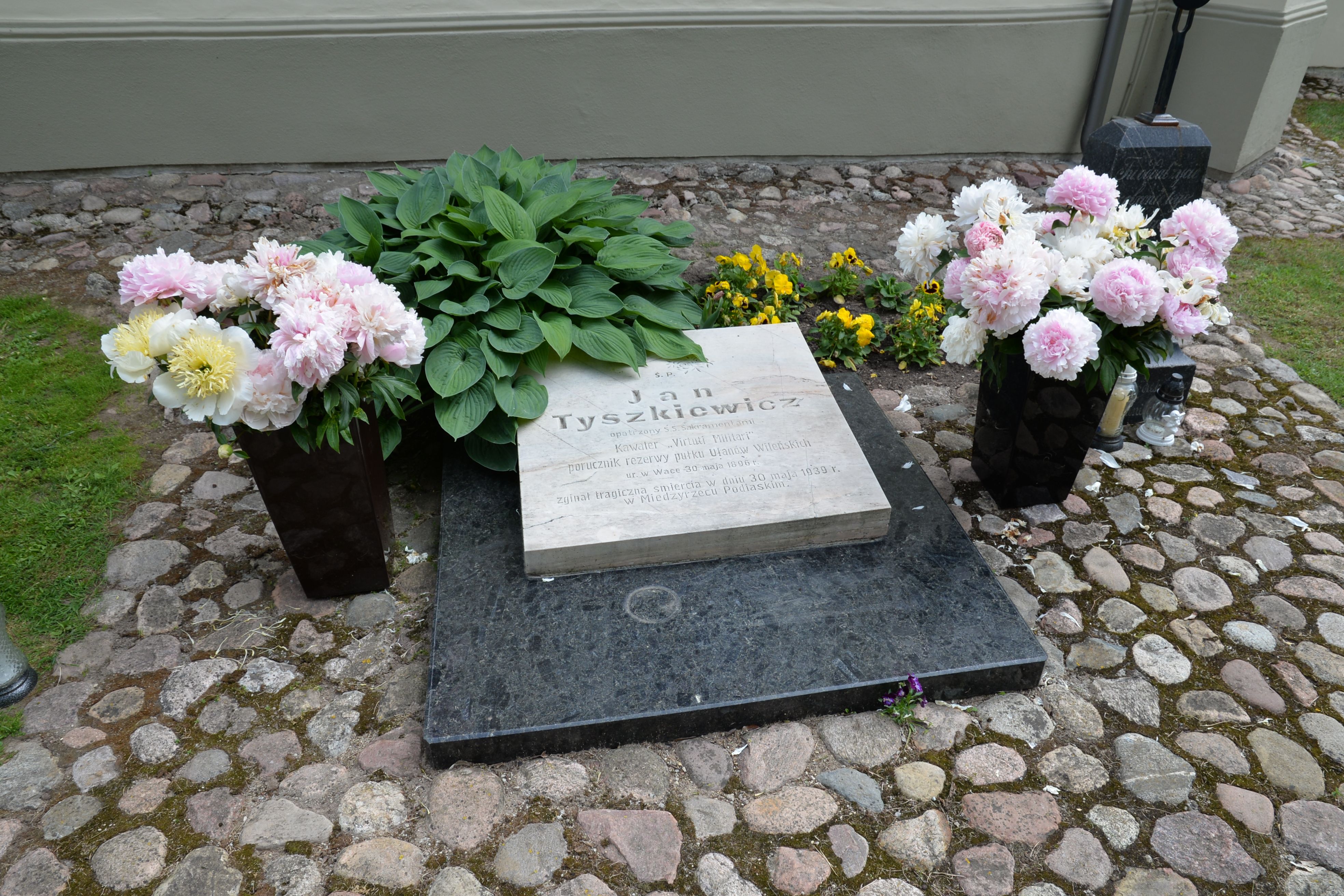 Pierre tombale de Jan Tyszkiewicz avec texte gravé, entourée de fleurs roses et blanches dans des vases, sur un chemin pavé.
