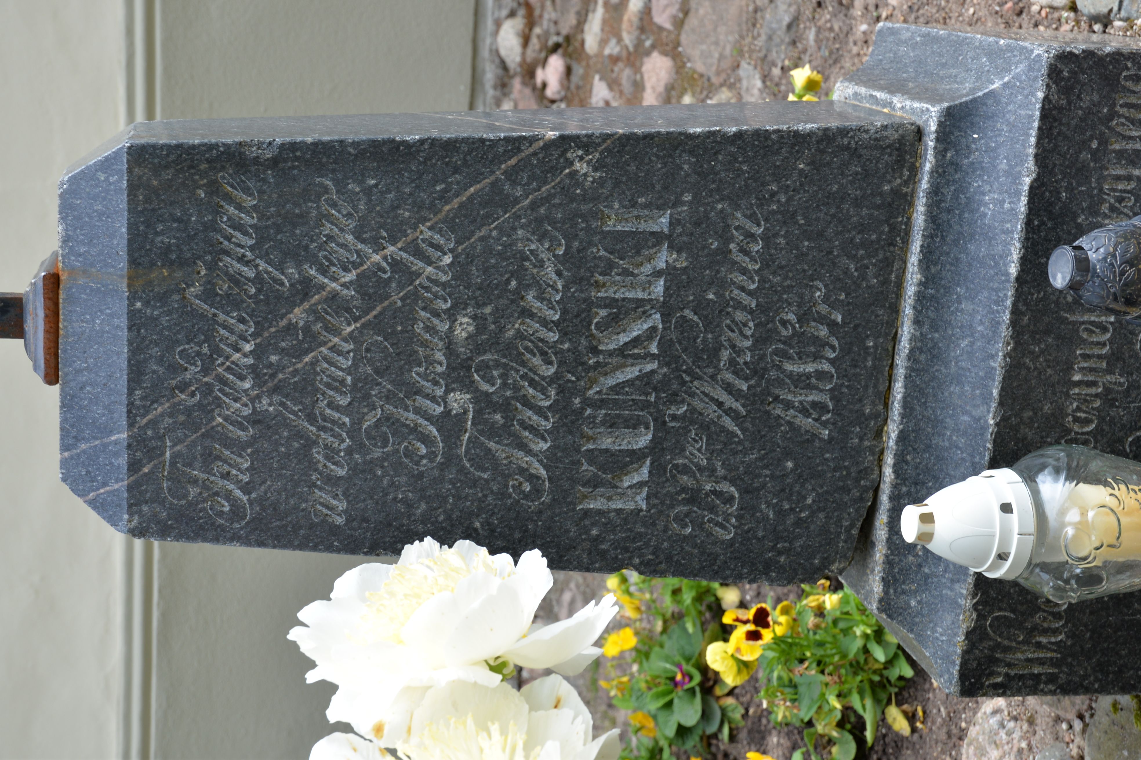 Pierre tombale de Tadeusz Kuński avec texte gravé en polonais, entourée de fleurs et de bougies. La pierre est sombre et usée, avec une croix au sommet.