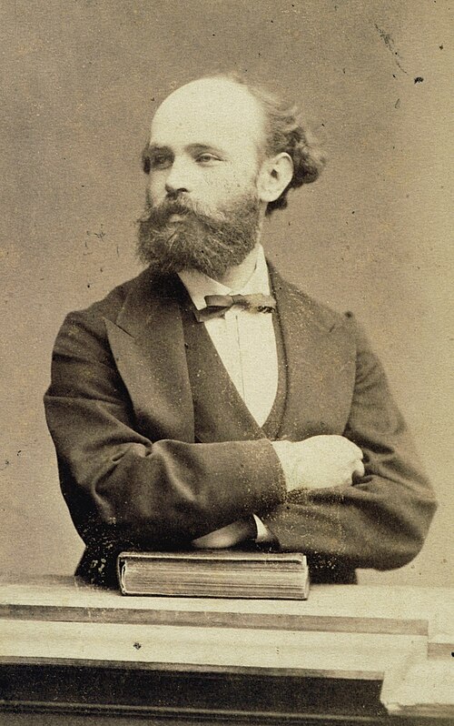 A sepia-toned portrait of a bearded man in a suit with crossed arms, standing behind a book on a table.