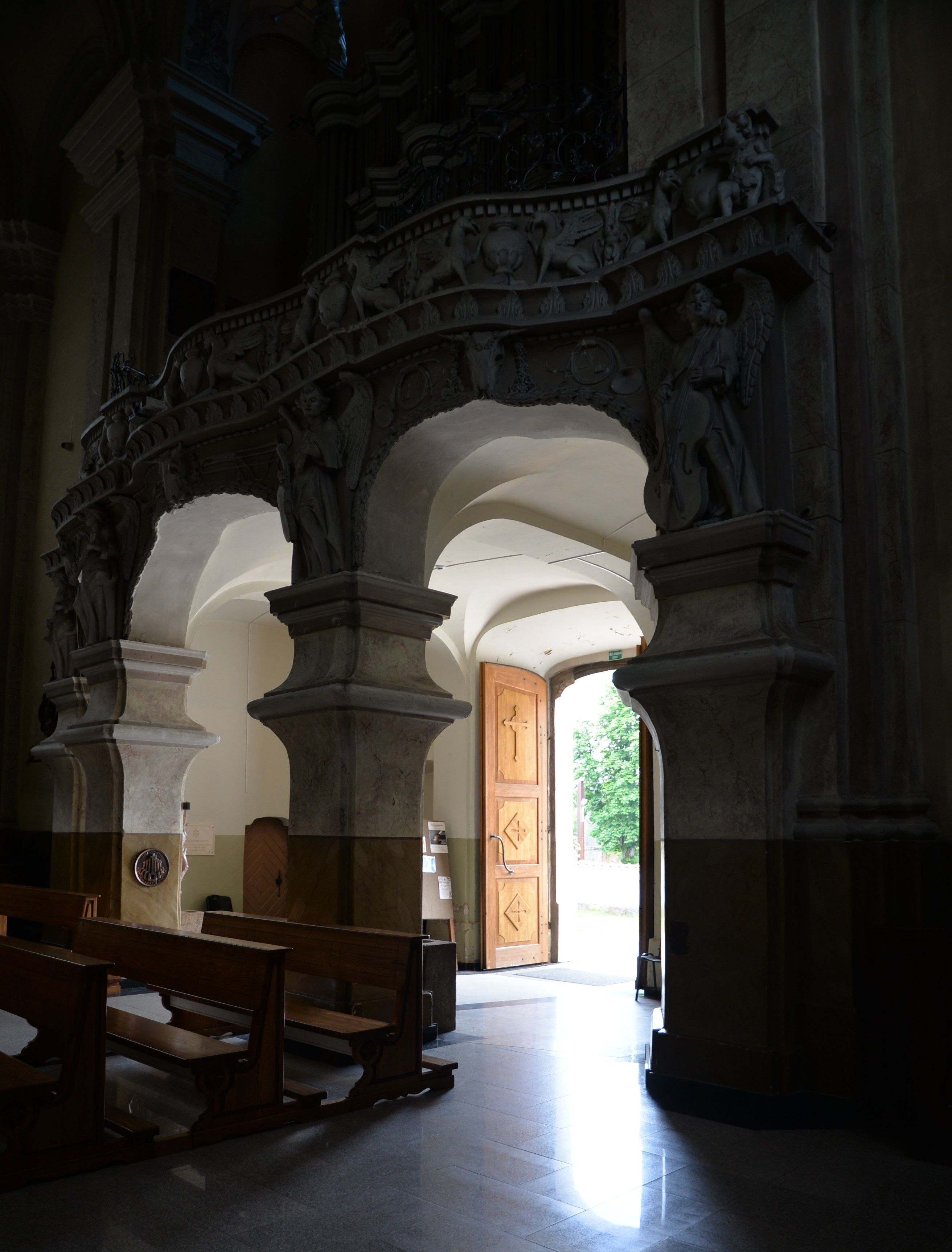 Intérieur de l'église de la Visitation de la Bienheureuse Vierge Marie à Szydłów, montrant des arcs ornés de sculptures d'anges et des bancs en bois. La lumière entre par une porte en bois ouverte.