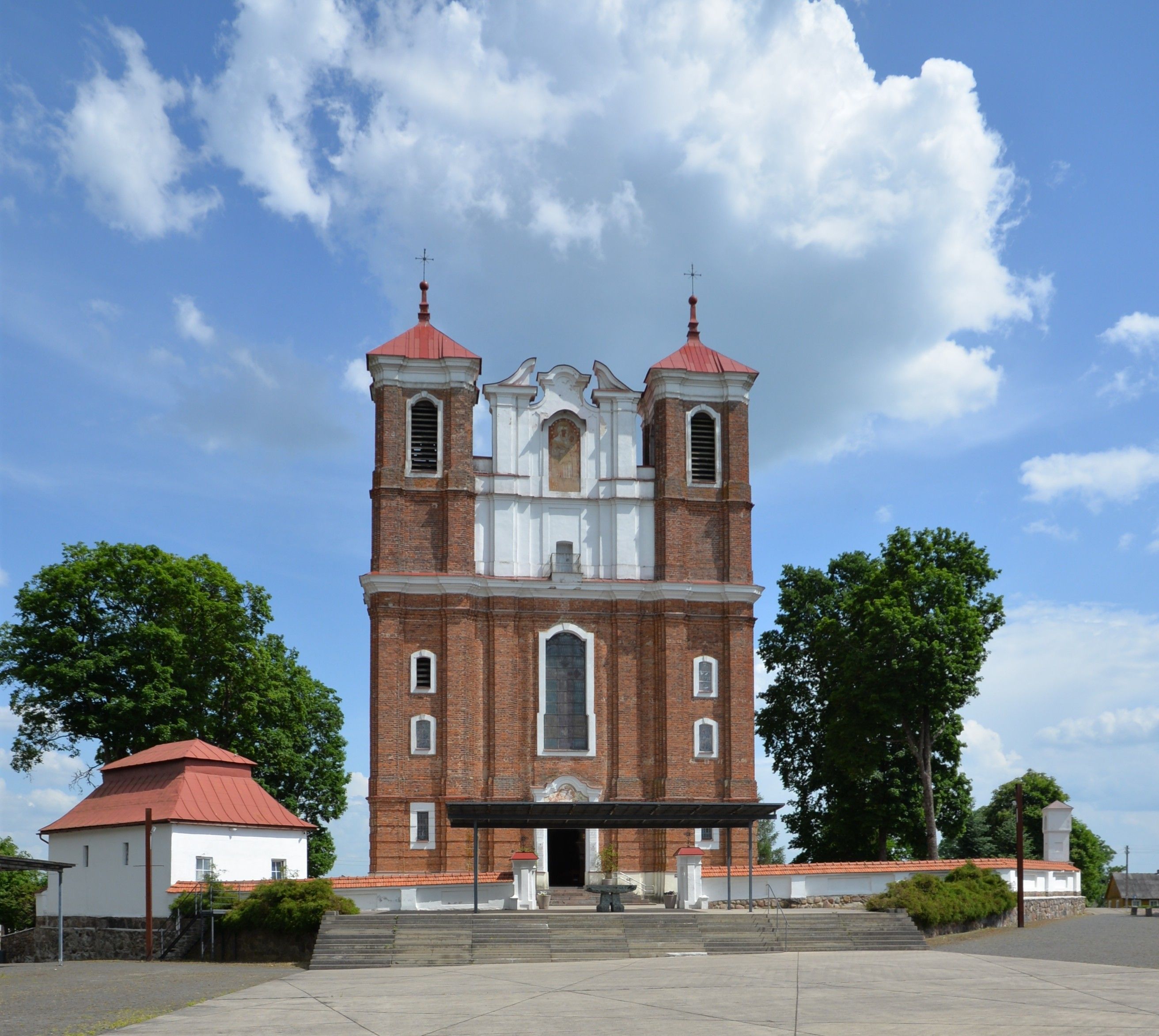 L'église de la Visitation de la Bienheureuse Vierge Marie à Szydłów, Lituanie, présente une façade en briques rouges avec deux tours carrées et un fronton central. Des arbres et un petit bâtiment au toit rouge sont visibles à proximité.