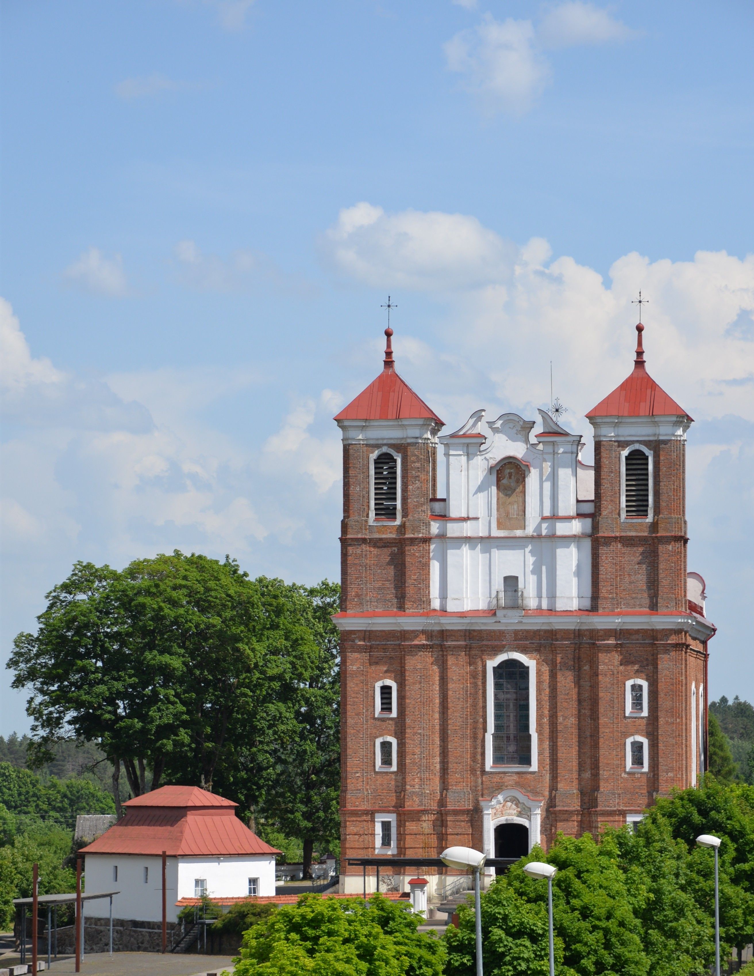L'église de la Visitation de la Bienheureuse Vierge Marie à Szydłów, Lituanie, présente une façade en briques rouges avec deux tours carrées et un haut fronton. Des arbres et un petit bâtiment au toit rouge sont visibles à proximité.