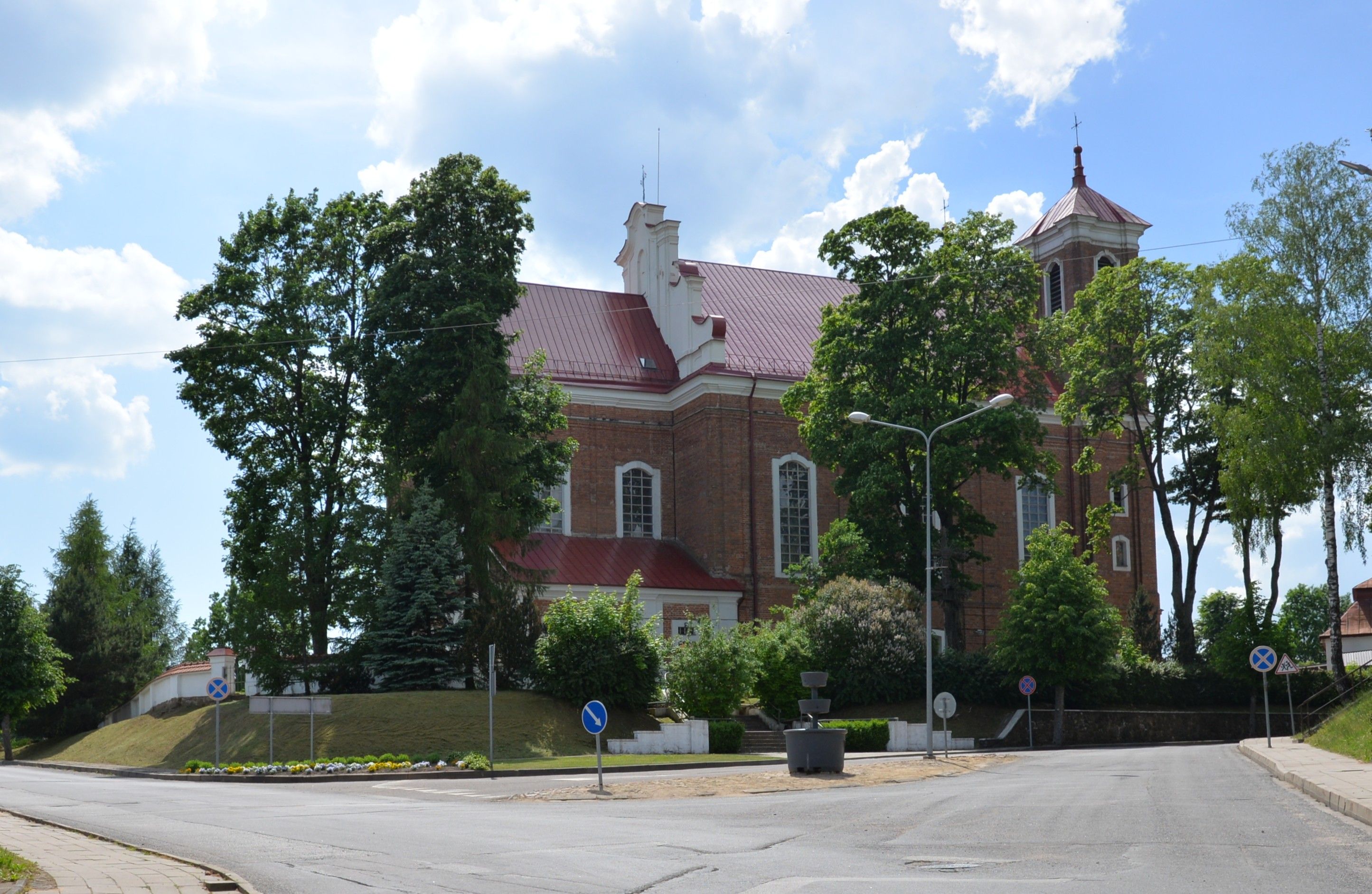 Église de la Visitation de la Bienheureuse Vierge Marie à Szydłów, Lituanie, avec des murs en briques rouges, entourée d'arbres et une rue au premier plan.