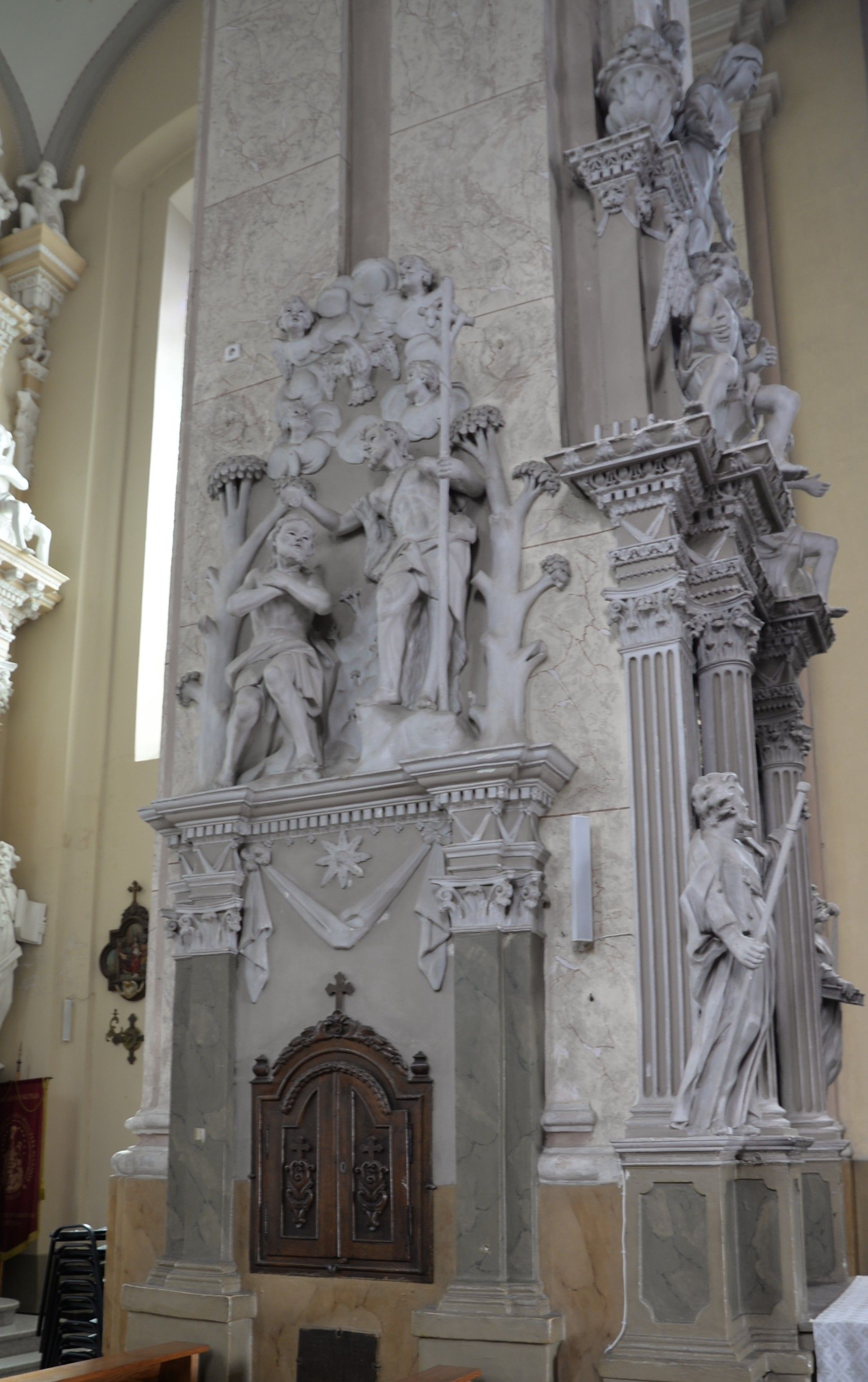 Intérieur de l'église de la Visitation de la Bienheureuse Vierge Marie à Szydłów, avec des figures en stuc baroques-classiques et des éléments architecturaux. Une porte en bois avec des sculptures ornées est visible en dessous.