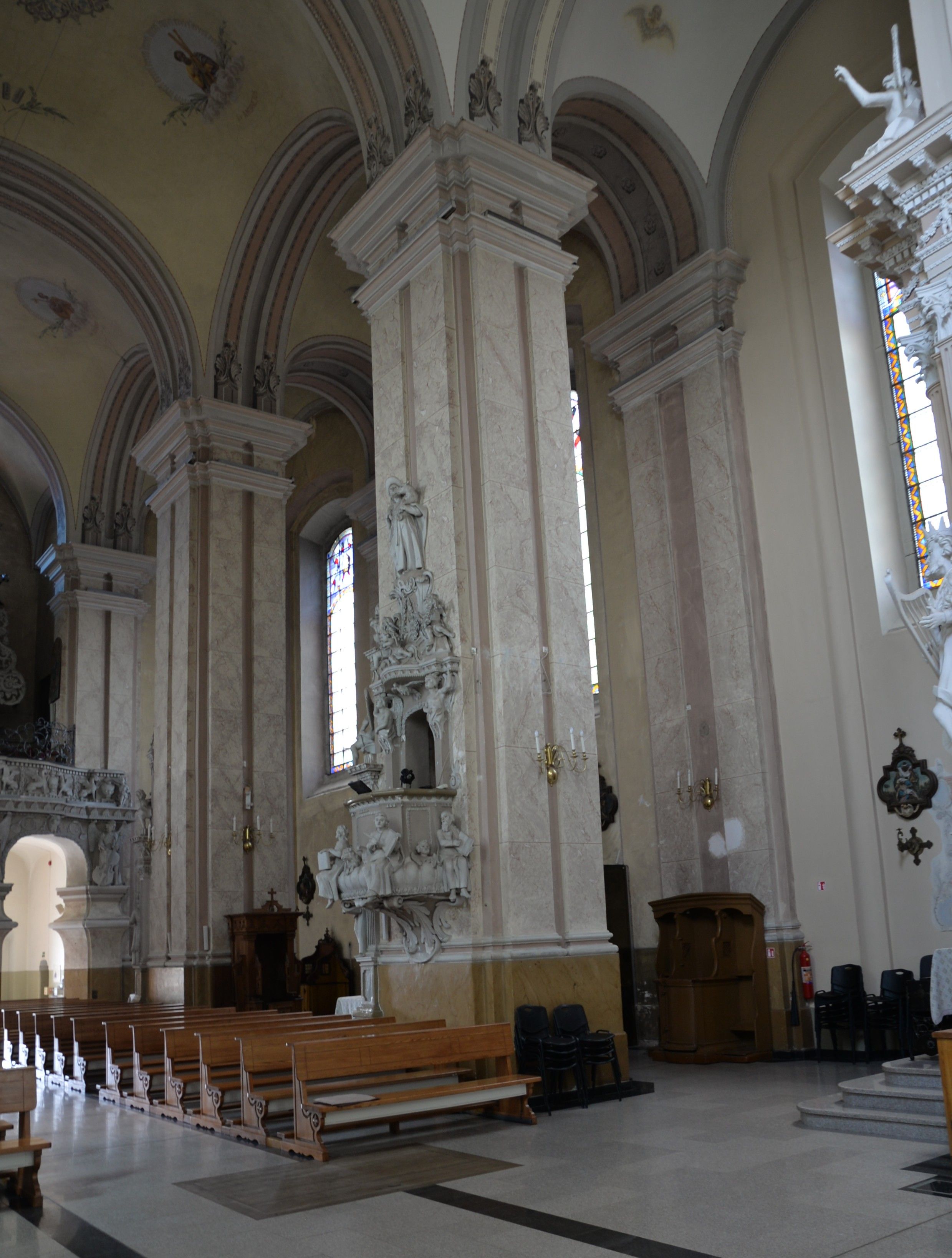 Intérieur de l'église de la Visitation de la Bienheureuse Vierge Marie à Szydłów, avec de hautes colonnes ornées de stuc, des bancs en bois et des vitraux.