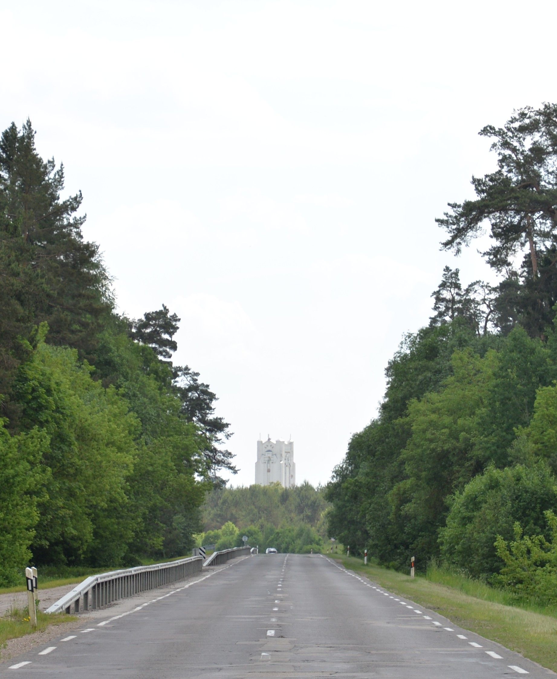 Дорога, що веде через ліс, з високою білою каплицею, видимою вдалині. Каплиця оточена деревами і виділяється на тлі неба.