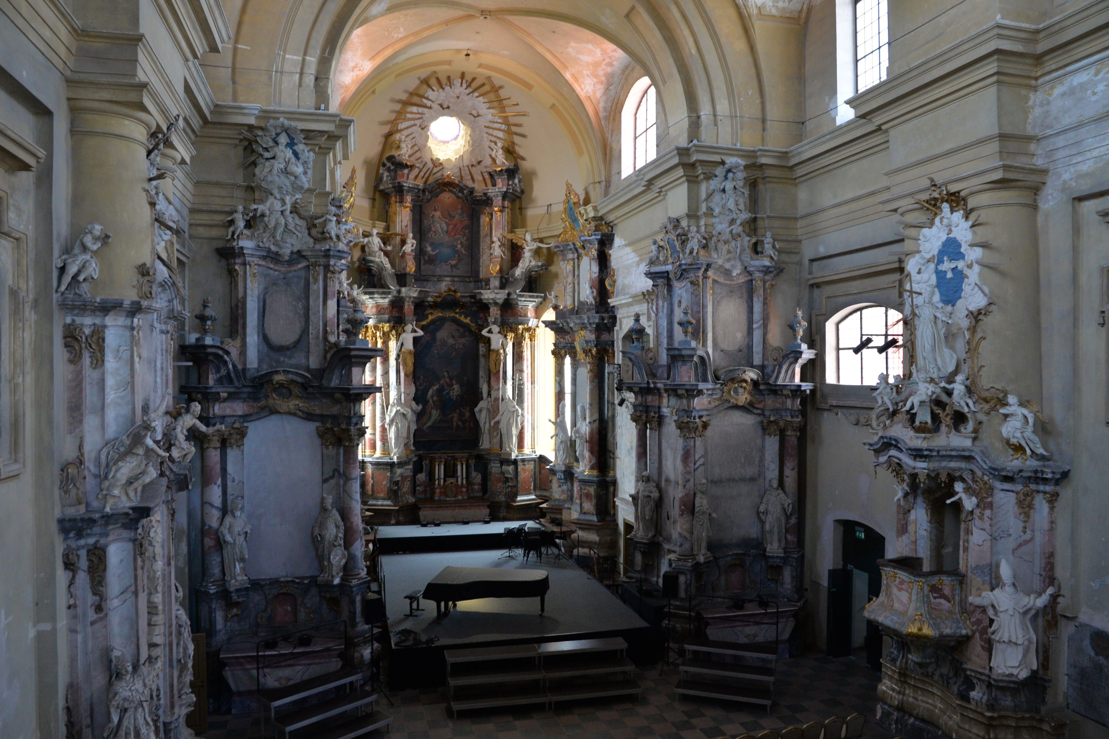 Interior of the Church of St. Catherine in Vilnius, featuring ornate baroque altars and sculptures. A grand piano is placed on a stage in front of the altar.