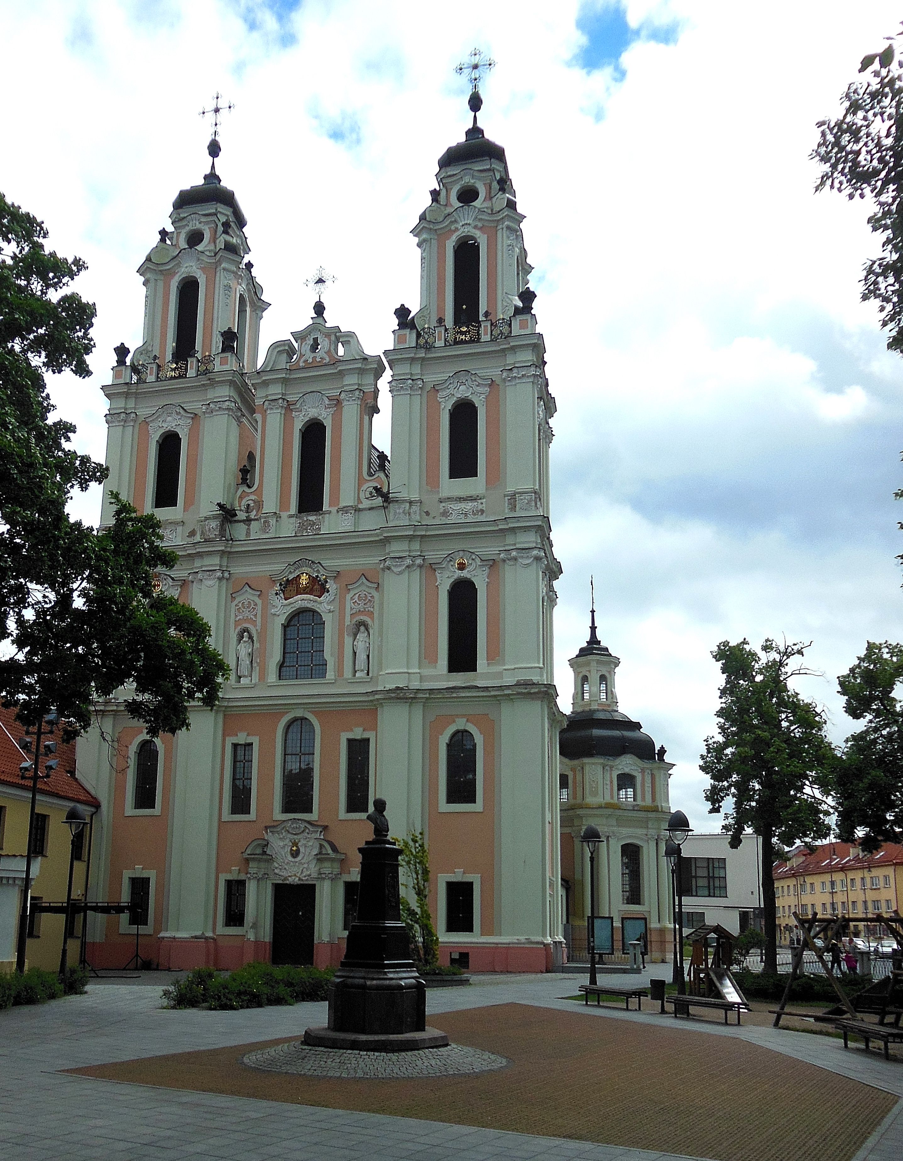 Facade of the Benedictine Church of St. Catherine in Vilnius with two towers and a central entrance. A statue stands in the foreground, surrounded by trees and benches.
