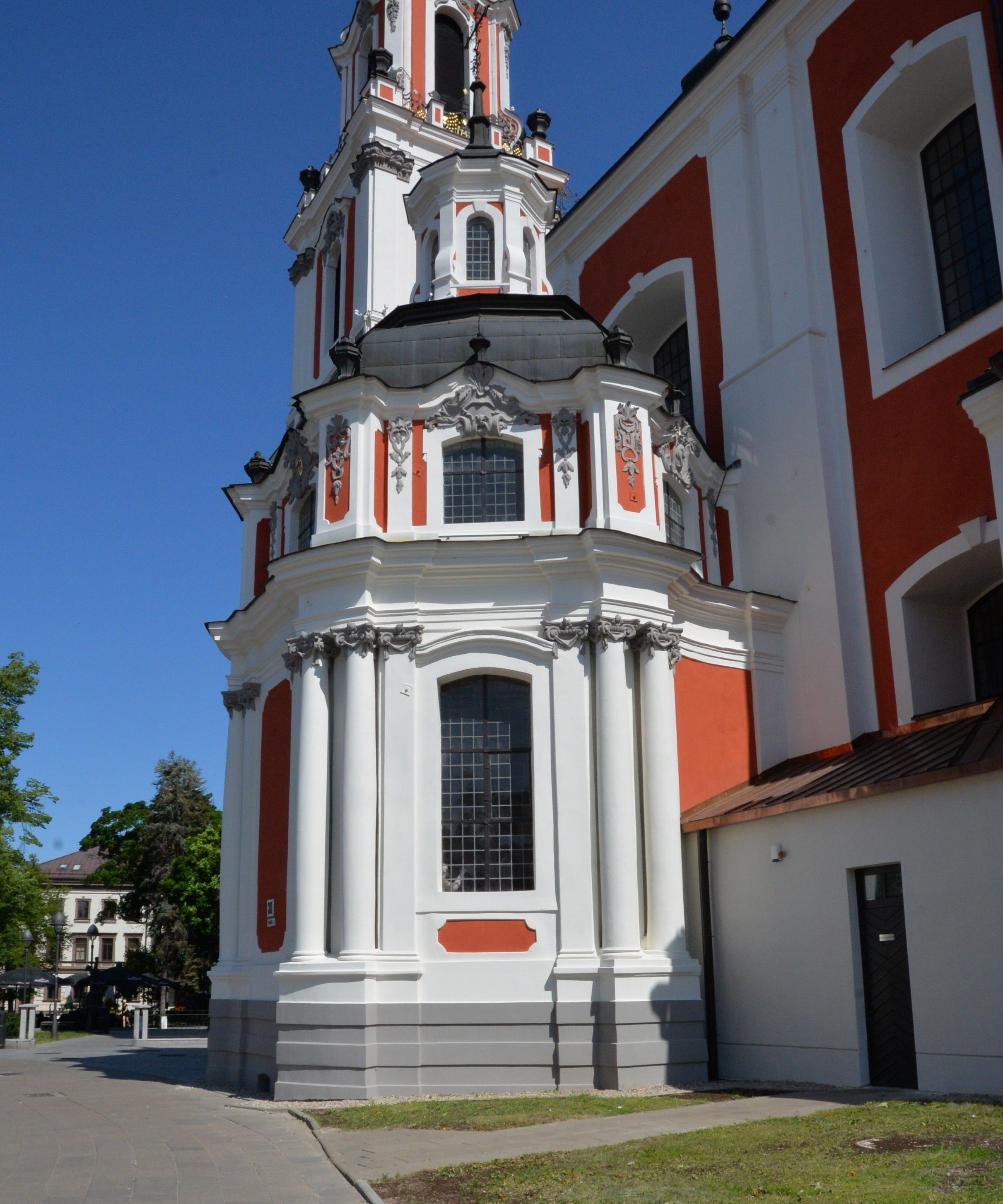 Facade of the Benedictine Church of St. Catherine in Vilnius, featuring a tall, ornate tower with red and white detailing, large windows, and decorative elements against a clear blue sky.