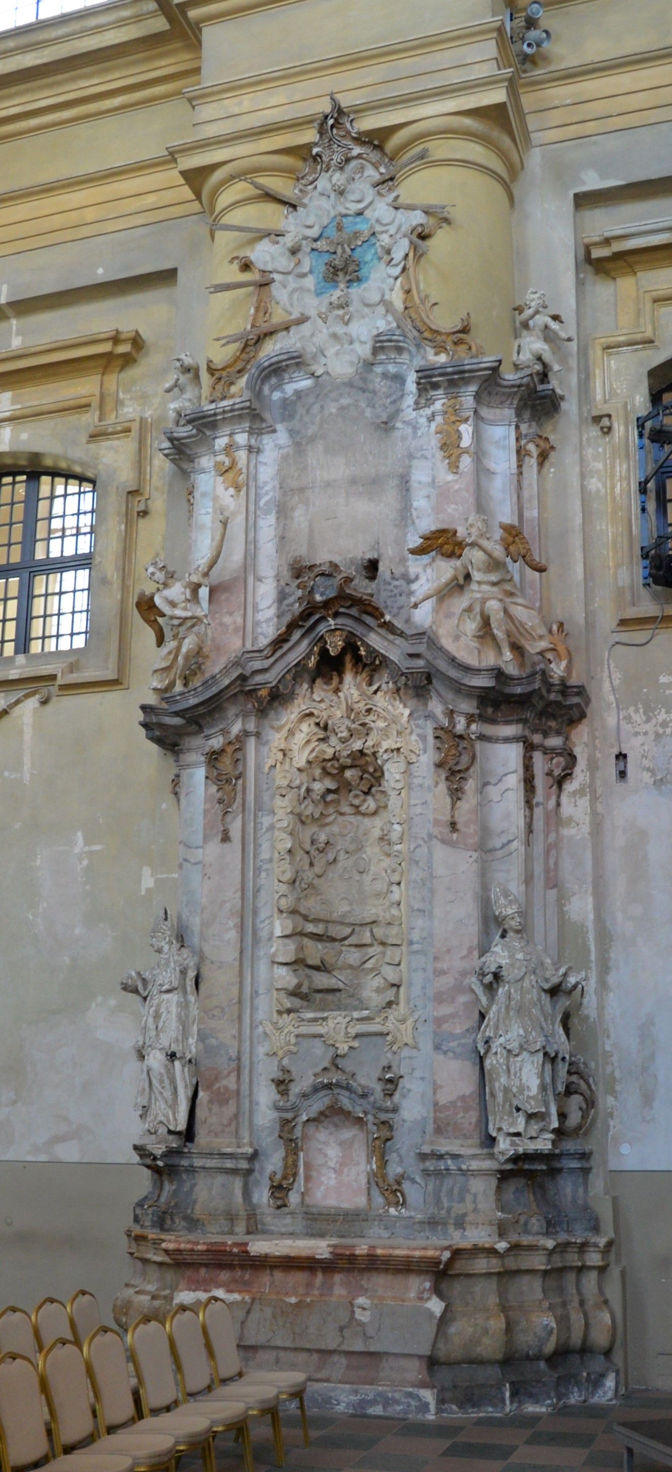 Interior of the Church of St. Catherine in Vilnius, featuring a richly decorated baroque altar with sculptures of angels and saints. The altar is flanked by columns, and chairs are arranged in front.