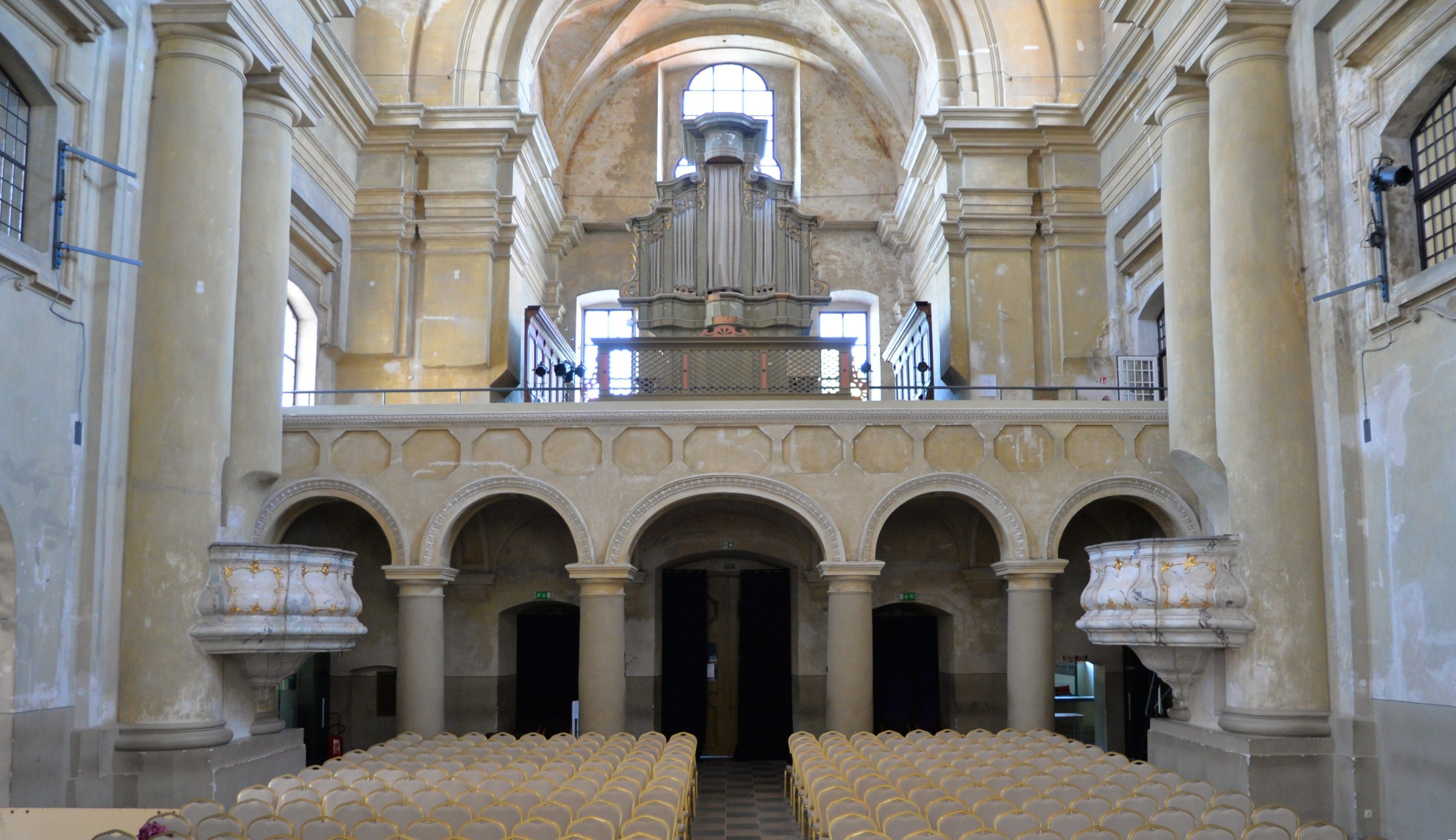 Interior of the Church of St. Catherine in Vilnius, featuring a view of the organ loft with a large pipe organ. The space is adorned with Tuscan columns and arches, and rows of chairs are arranged for concerts.