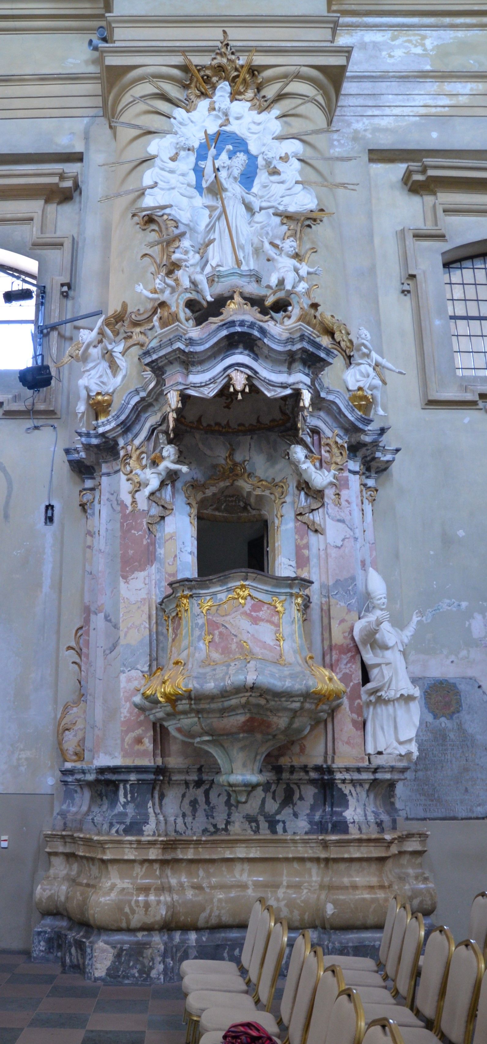 Baroque pulpit in the Church of St. Catherine in Vilnius, adorned with gold and marble details, featuring sculptures of saints and angels. Rows of chairs are visible in the foreground.