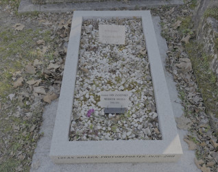 Grób Lucasa Dolegi z niską ramą wypełnioną kamieniami. Inskrypcje na ramie i dwie tabliczki na kamieniach. Odręczna płytka z rysunkiem karykatury człowieka w spiczastej czapce.