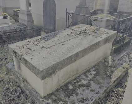 Kamienna tumba z płaską nakrywą i wypukłym krzyżem, otoczona innymi grobami. Na bokach i wierzchu tumby znajdują się inskrypcje. Wokół podstawy widoczny jest mech i opadłe liście.