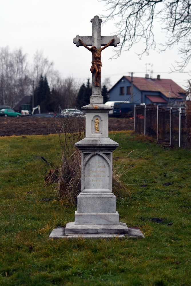 Roadside cross in grassy area with stone base and metal statue of Christ. Cross surrounded by a simple iron fence, with houses and trees in the background.