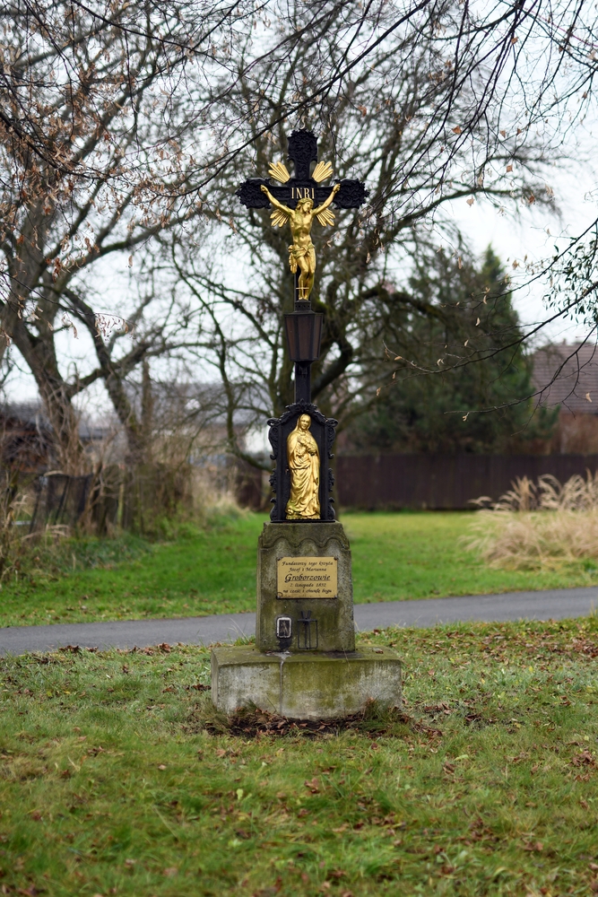 An iron cross with a golden figure of Christ and the inscription INRI, set on a stone base with a plaque in Polish. Surrounded by grass and trees, it is located in the Czech part of Těšín Silesia.