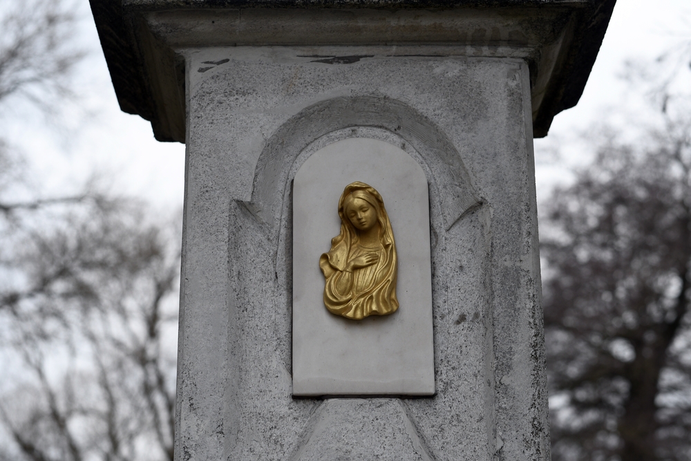 A stone wayside shrine in the Czech part of Těšín Silesia with a golden relief of the Virgin Mary. The chapel is surrounded by leafless trees, indicating a winter setting.