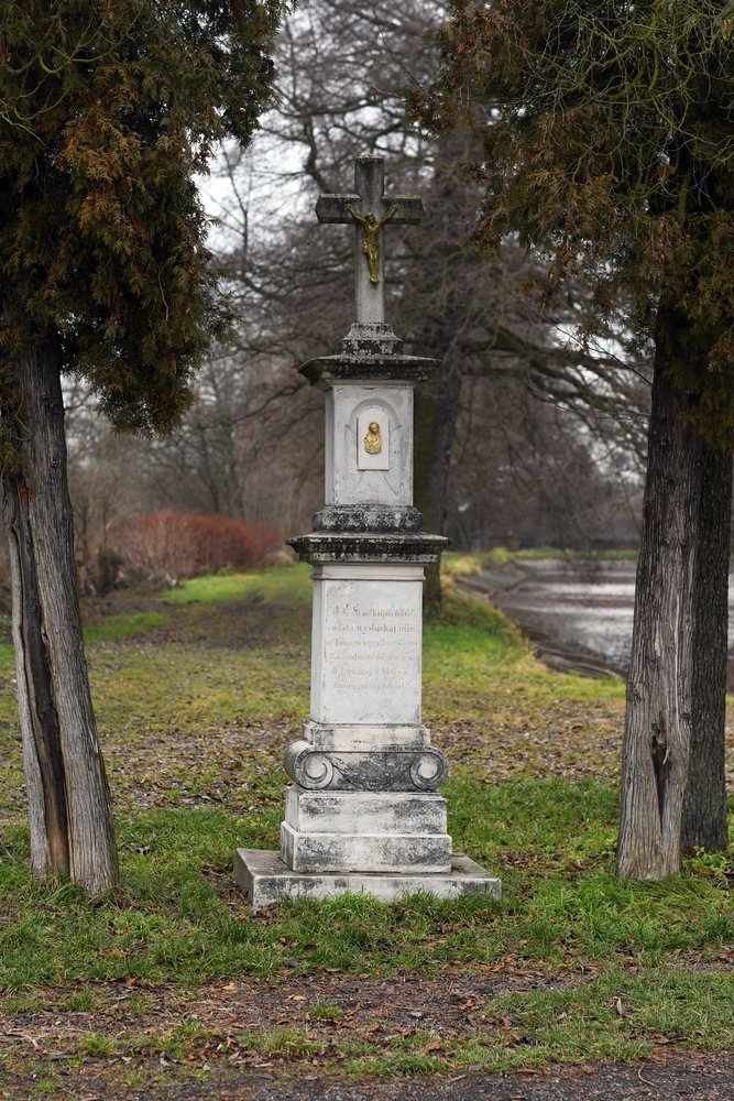 A stone roadside cross in a grassy area, surrounded by two trees. The cross has a small crucifix and an inscription on the plinth. Bare trees and a path can be seen in the background.