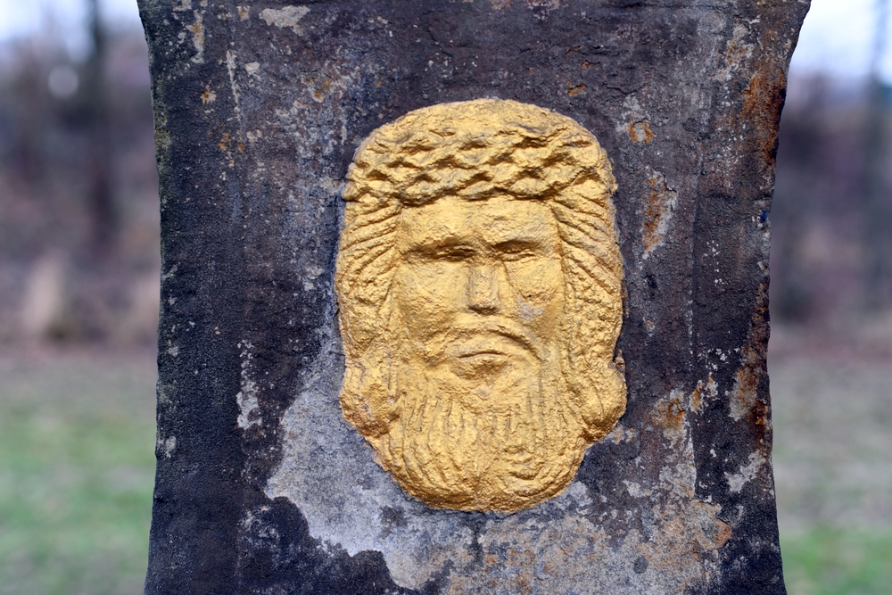 Destroyed stone relief depicting the face of a bearded man with a crown of thorns, painted in gold, on a dark textured background.