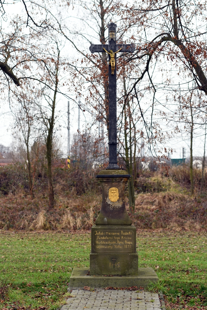 An iron cross with a brass figure of Christ on a stone plinth, surrounded by bare trees. The inscription reads: 'Jakób and Marianna Radecki, Founders of this Cross, To the honour and glory of the Lord God, erected Year 1871.'