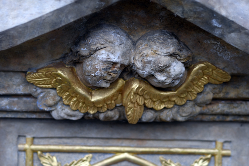 Two stone heads of cherubs with golden wings, part of a religious monument in Těšín Silesia, Czech Republic. The cherubs are weathered, with delicate facial features and detailed wings.