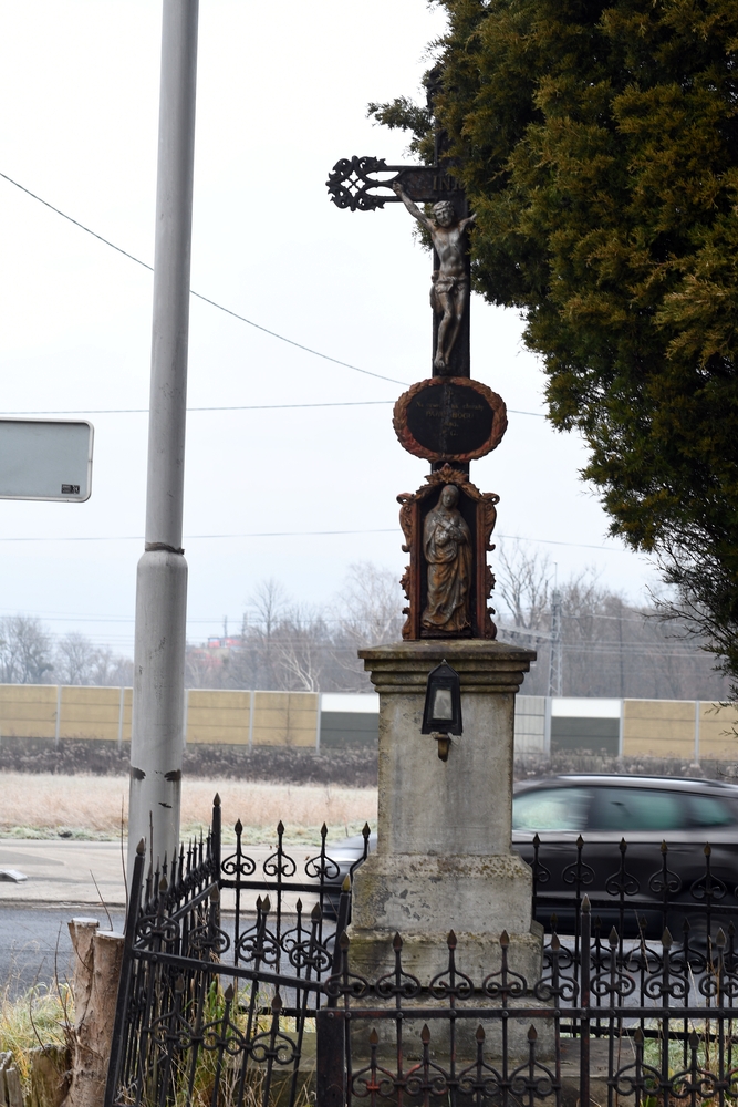 A wayside cross on the Czech side of Těšín Silesia, with a crucifix and a statue of the Virgin Mary below. Surrounded by an iron fence, it stands by the road with cars passing by.