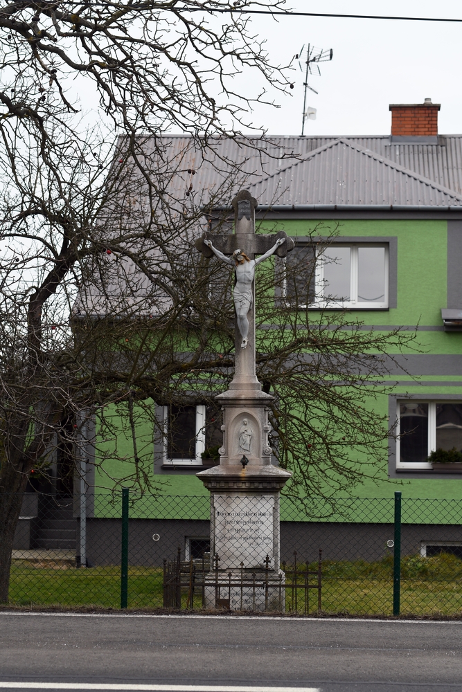 Roadside cross in front of a green house with metal fencing and bare trees. The cross has a statue of Christ and a small figure in a niche on a plinth.