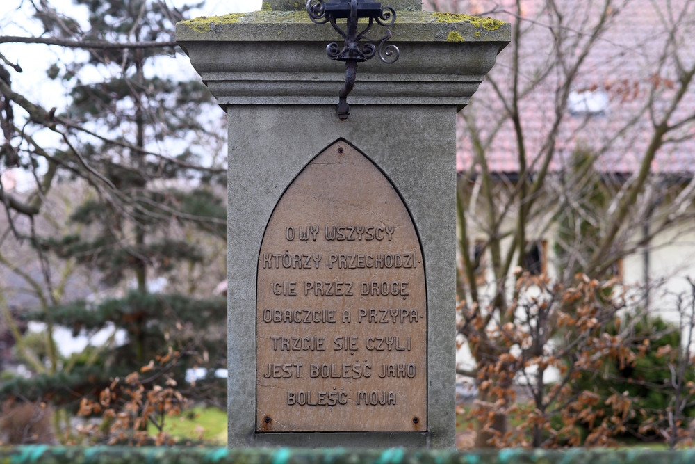 Close-up of a stone roadside cross with an inscription: 'O all of you who pass by on the road, observe and look, that is, sorrow as my sorrow.' Cross surrounded by trees, house visible in background.
