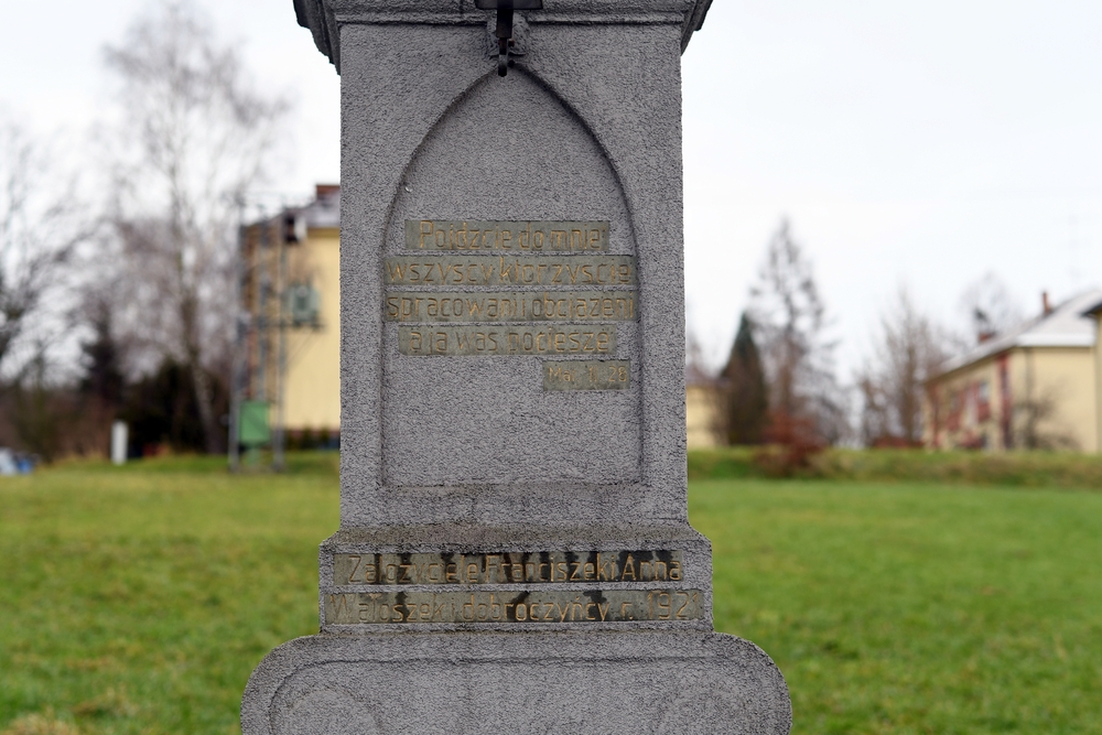 A stone cross plinth with inscriptions in Polish, placed on a grassy area with buildings in the background. The text reads: 'Come unto me, all ye that labour and are heavy laden, and I will comfort you.'