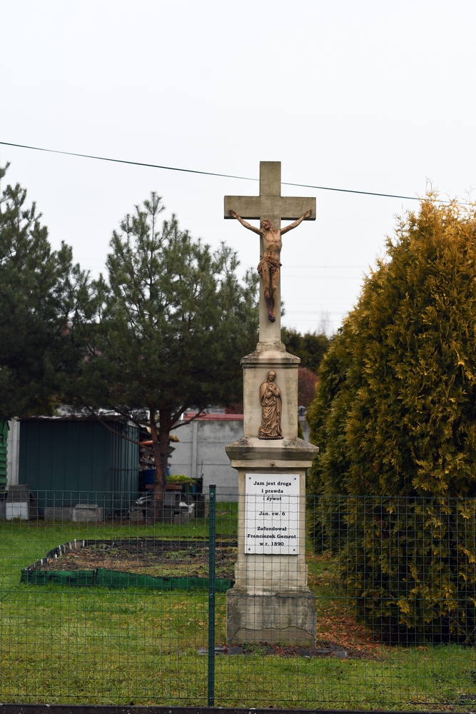Roadside cross in a grassy area, surrounded by a metal fence. Stone cross with crucifix and plaque with an inscription in Polish quoting John 14:6 and mentioning the founder, Franciszek Czernot, dated 1890.