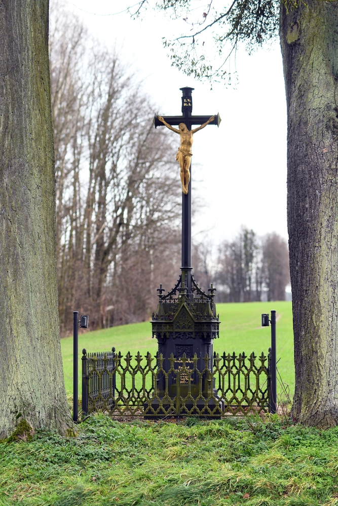 An iron cross with a golden figure of Christ stands between two trees, surrounded by an ornate wrought iron fence. The base has a neo-Gothic design with gilded inscriptions.