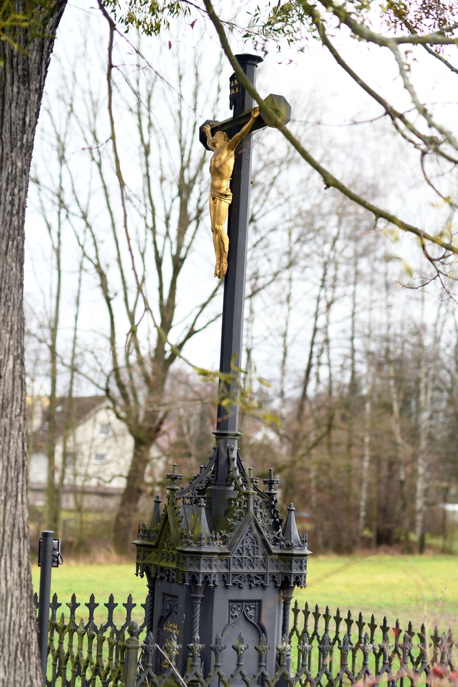 An iron cross with a golden figure of Christ stands in a fenced area, surrounded by trees. The base has neo-Gothic elements with intricate detailing.