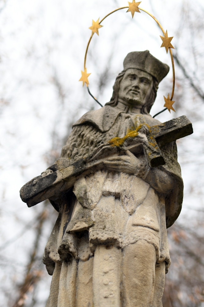 Stone statue of St John of Nepomuk holding a cross, with a halo of five stars. The figure is detailed, in traditional clerical garb, against a background of fuzzy, leafless trees.