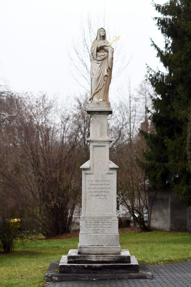 A stone statue of the Virgin Mary holding a cross stands on a high plinth with inscriptions, surrounded by trees and grass in the park.