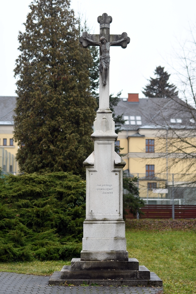 A stone cross with a figure of Christ stands on a plinth in a grassy area. The inscription reads: 'I am the way, the truth and the life' (John 14:6). Trees and a building are visible in the background.